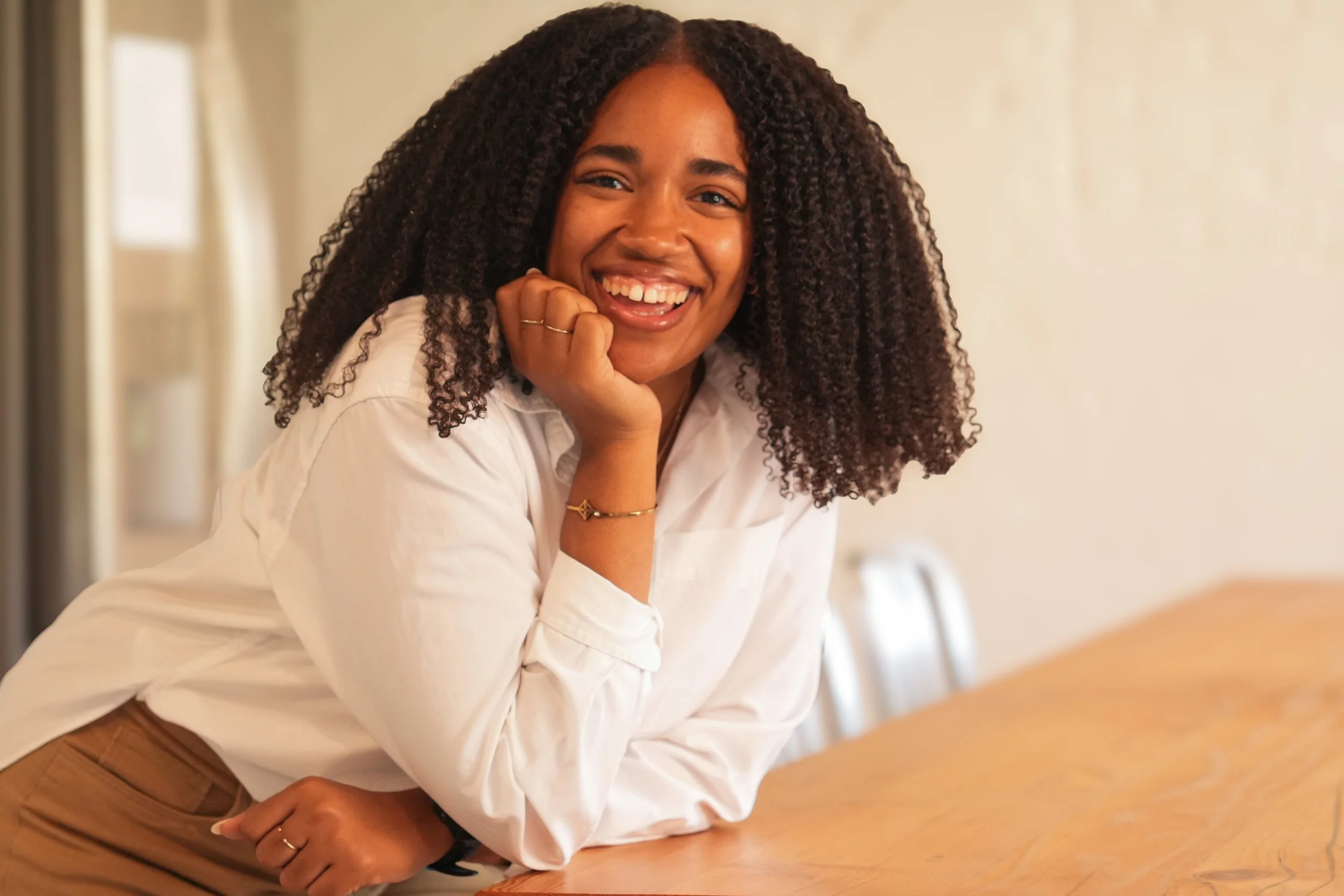 A smiling woman with curly hair wearing a white blouse leaning on a wooden table.