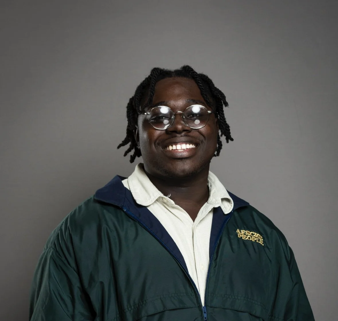 Smiling young man with glasses, dreadlocks, wearing a beige collared shirt and a dark green jacket with a logo.