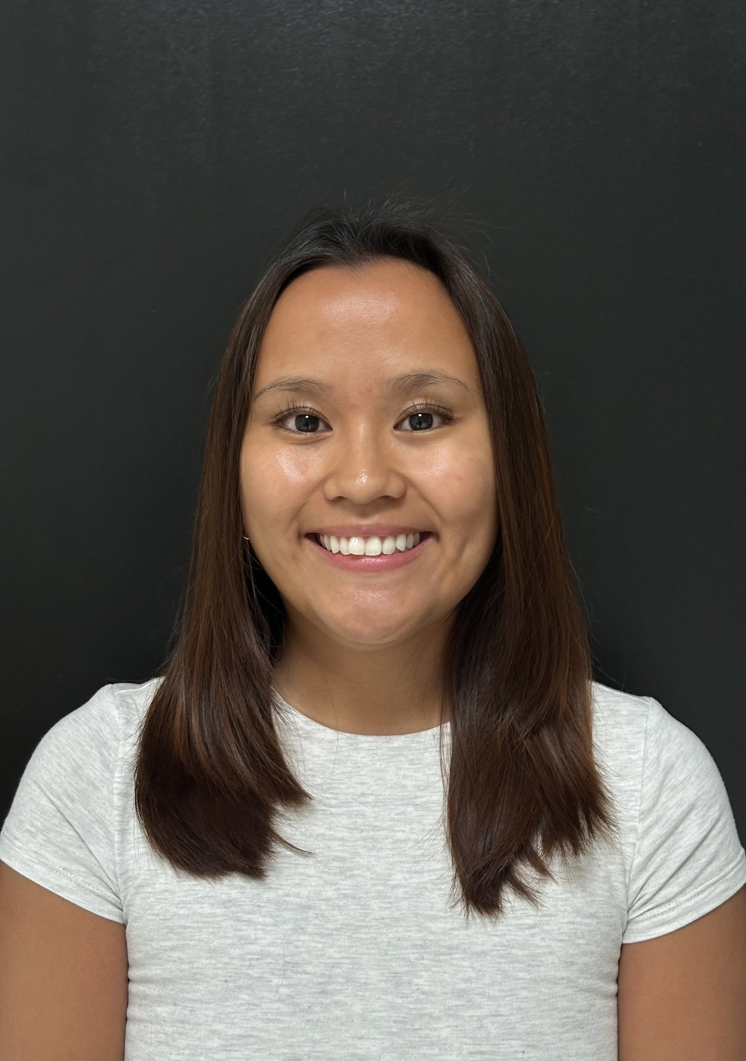 A young woman with shoulder-length brown hair, smiling, wearing a light-colored t-shirt, against a plain dark background.