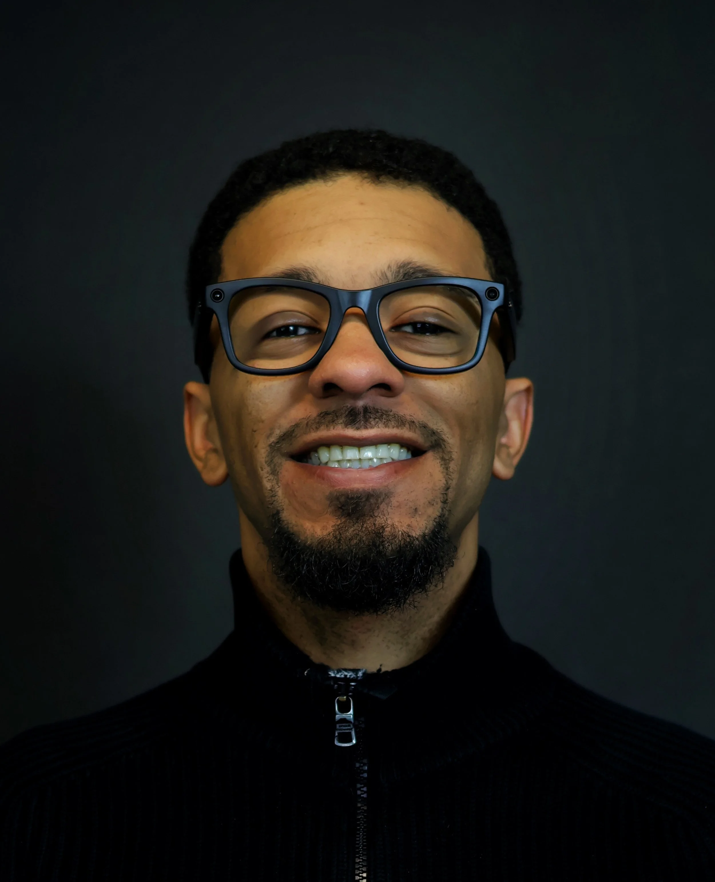 Close-up of a smiling man with glasses, dark hair, and a short beard wearing a black zip-up sweater against a dark background.