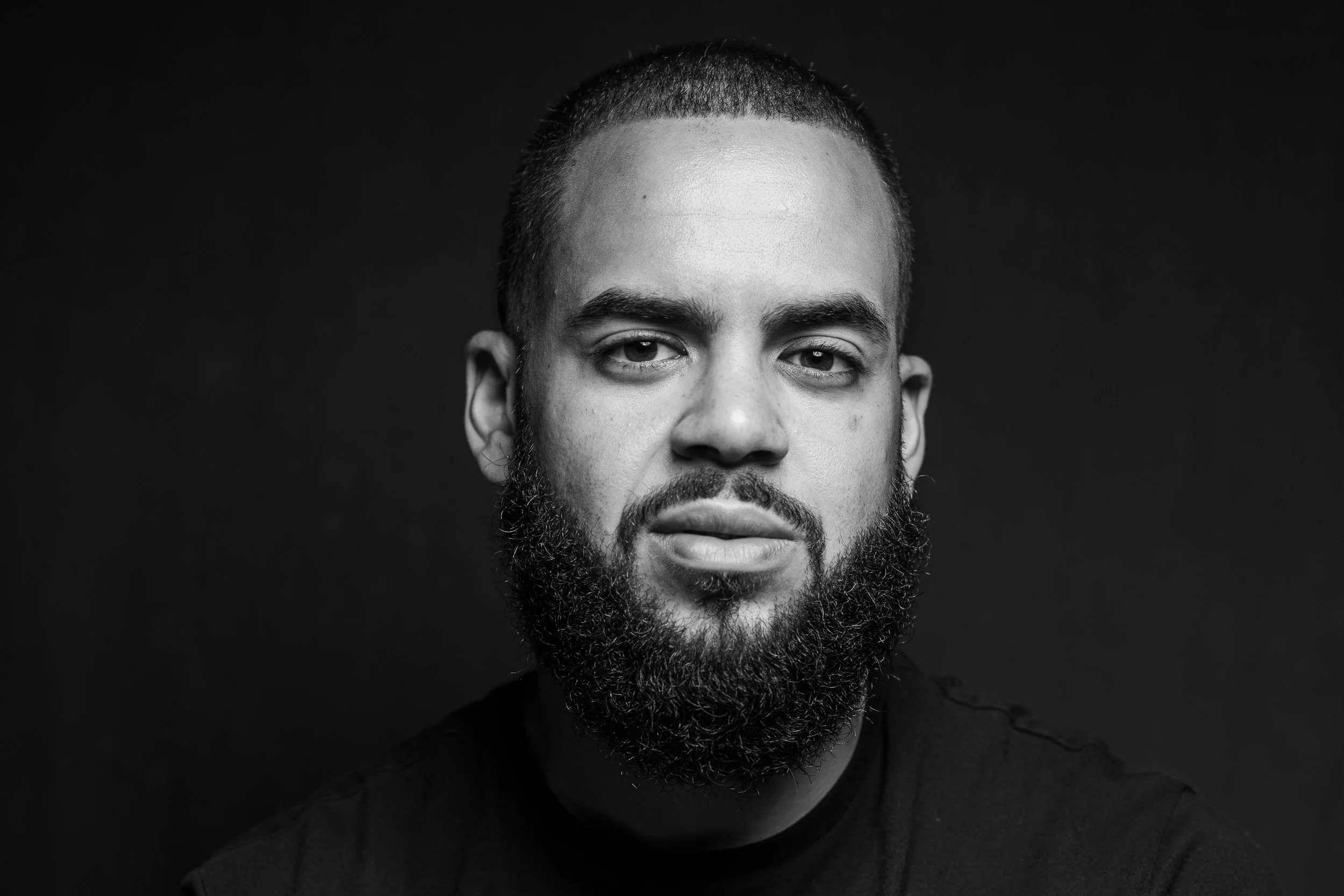 Black and white portrait of a man with a beard, short hair, and a serious expression, wearing a dark shirt against a dark background.
