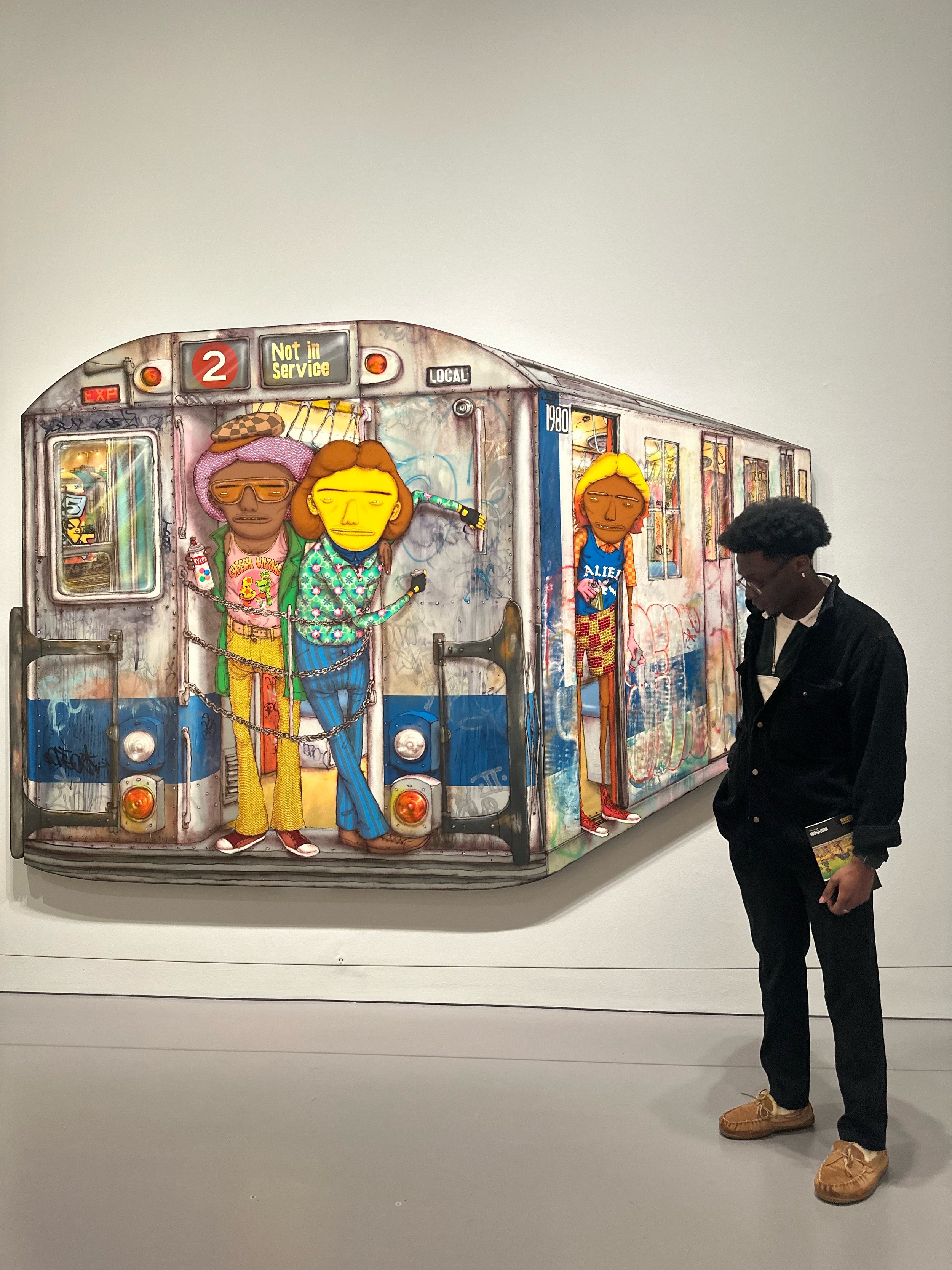 A person standing in front of a colorful graffiti-style artwork of a subway train with cartoon characters painted on it, inside an art gallery.