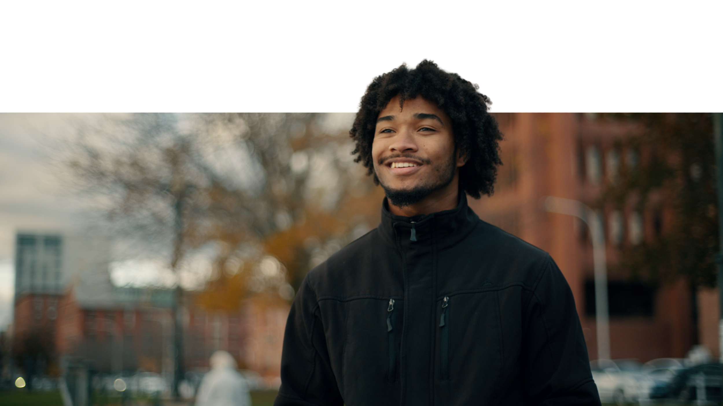Young man with curly hair and a black jacket smiling outdoors with blurred trees and buildings in the background.