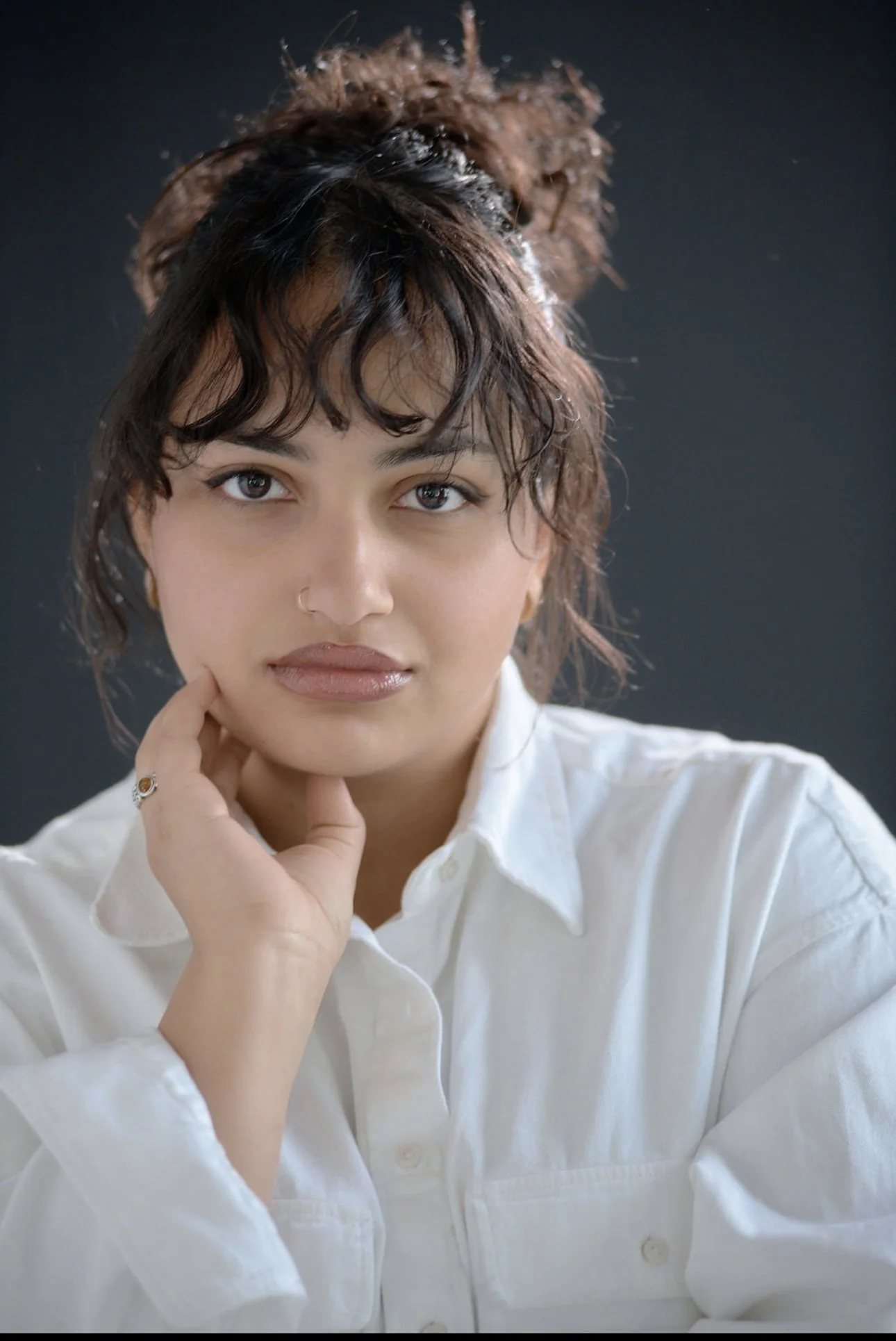 Portrait of a woman with dark curly hair in a messy bun, wearing a white shirt, against a dark background.