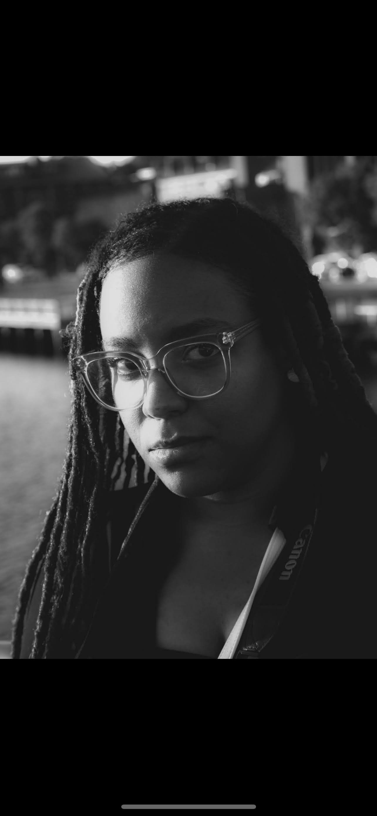 A black and white close-up photo of a woman with dreadlocks and eyeglasses, looking directly at the camera with a serious expression, with a blurred outdoor background.