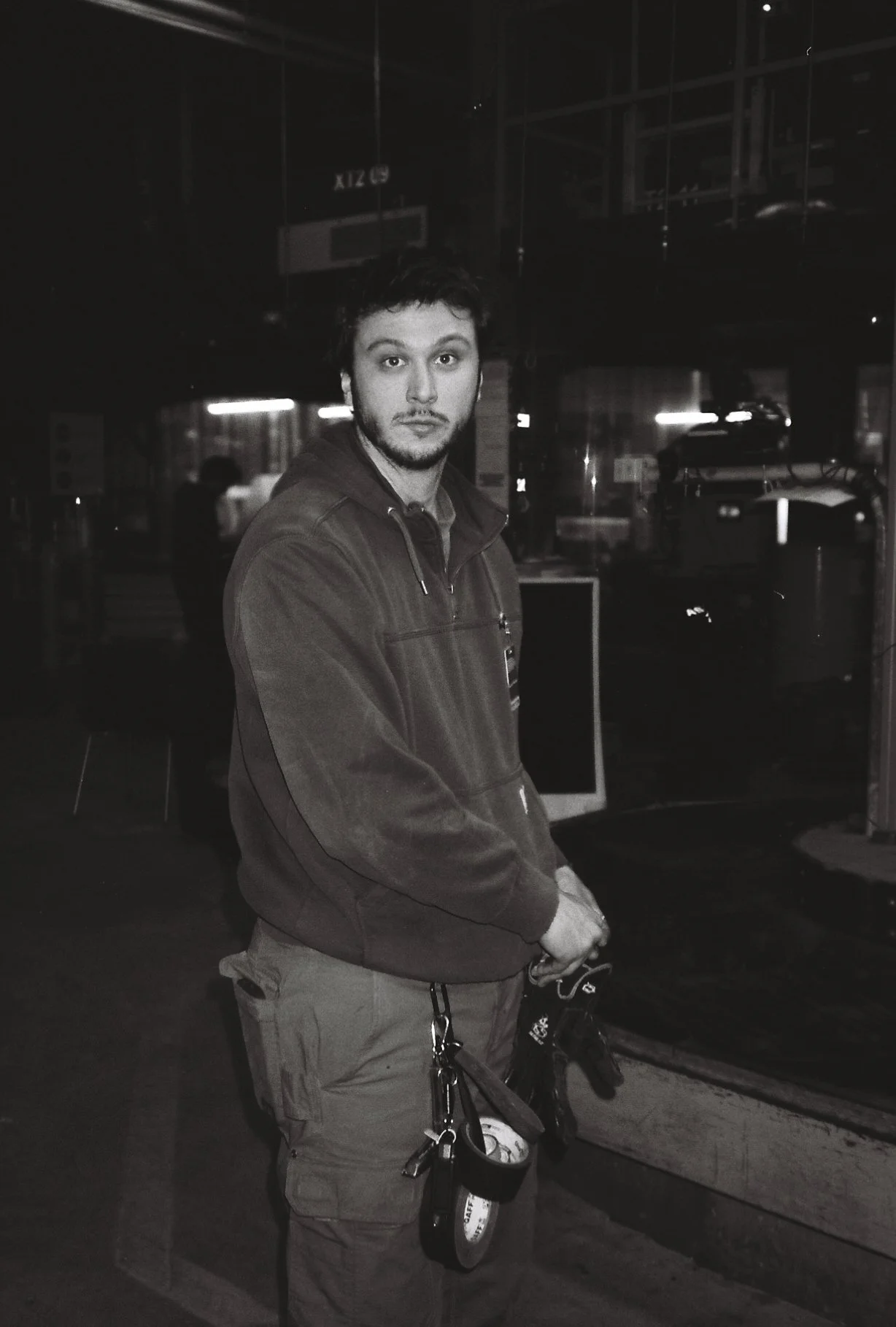 Young man with dark hair and beard wearing a hoodie and cargo pants, standing indoors at night, holding keys and a wristwatch, looking at the camera.