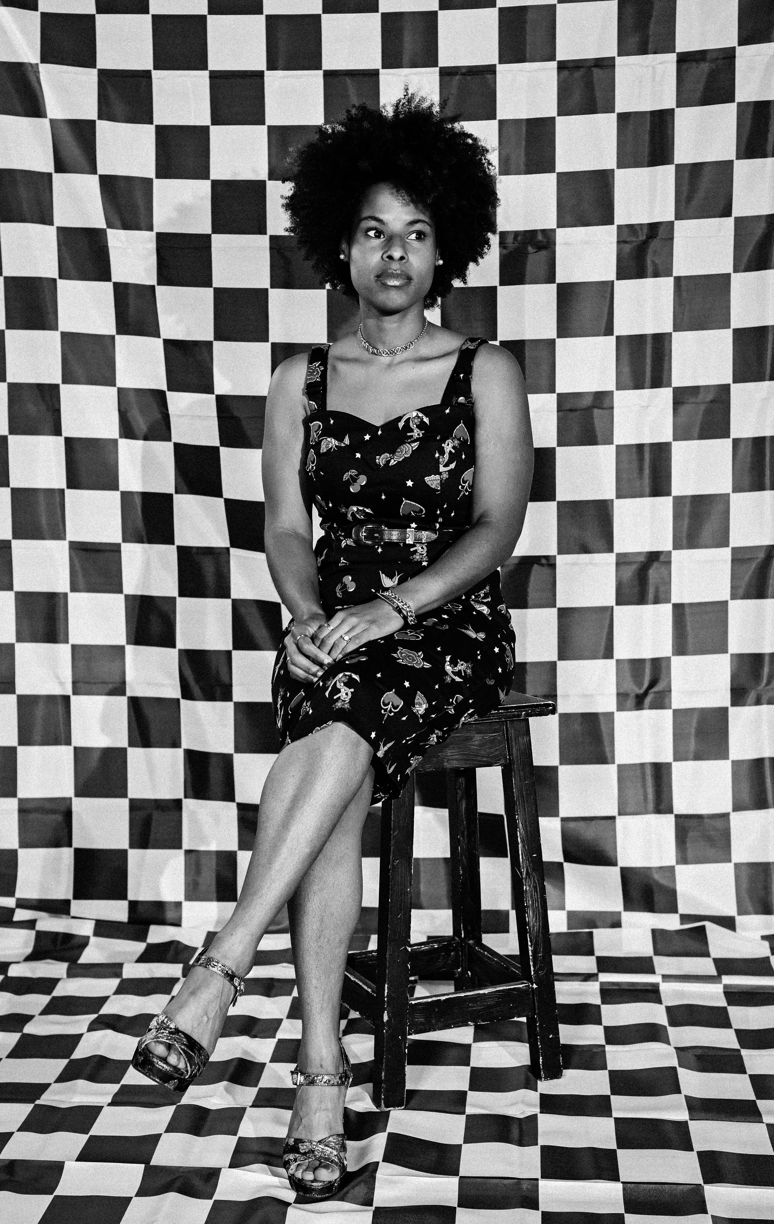 A woman with curly hair wearing a patterned dress, jewelry, and high heels sitting on a wooden stool in front of a checkered backdrop in black and white photography.