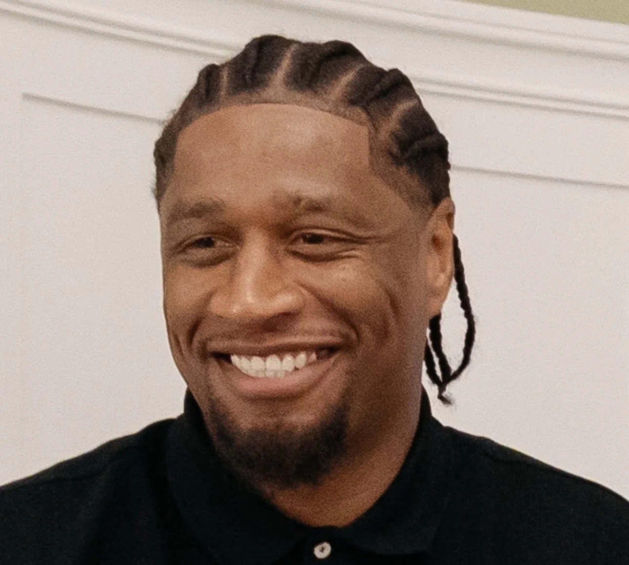 A smiling man with dreadlocks and a goatee, wearing a black collared shirt, in front of a plain white wall.
