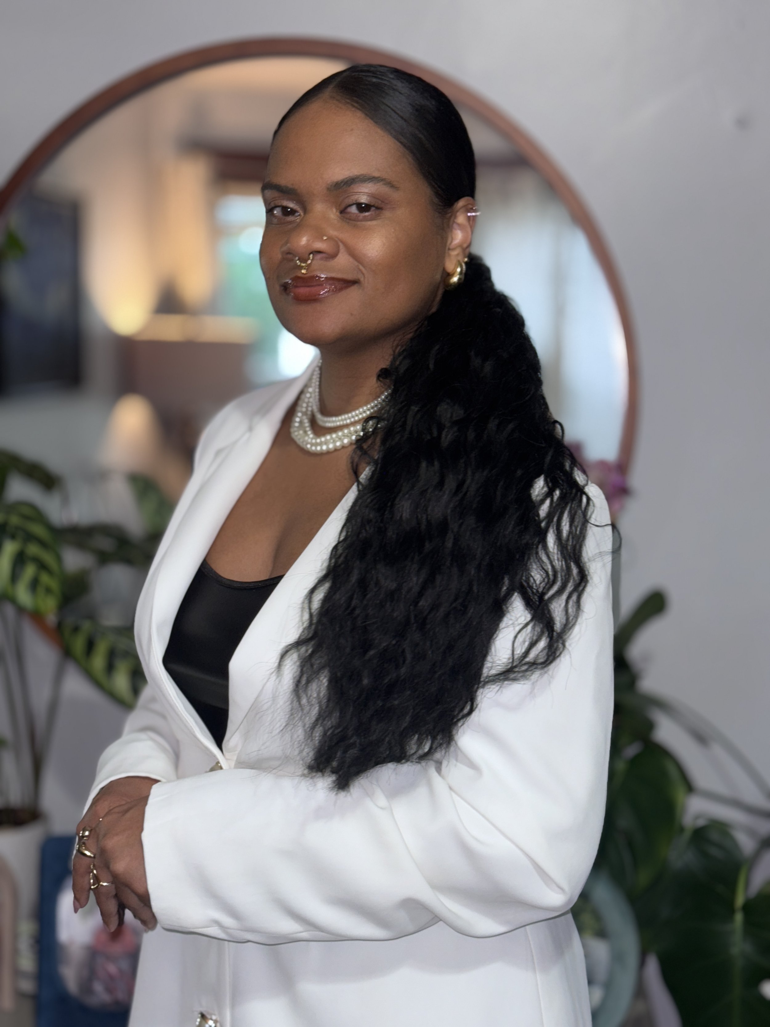 A woman with dark skin and long black wavy hair, wearing a white blazer, black top, pearl necklace, and gold jewelry, standing indoors in front of a mirror and plants.