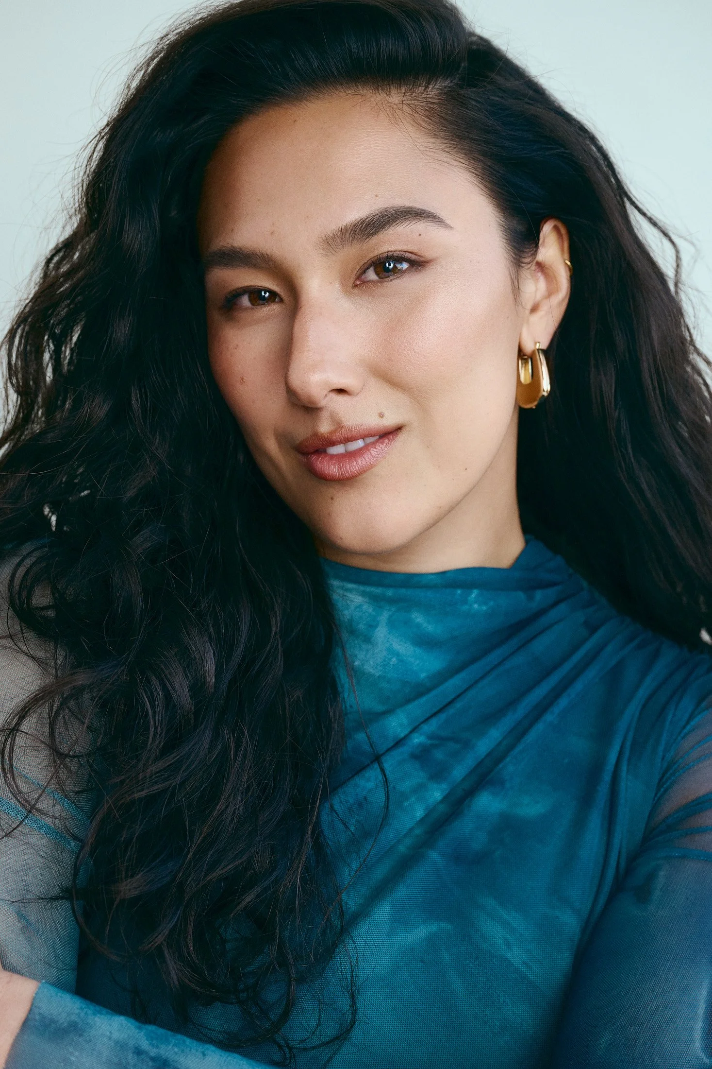 A woman with long dark wavy hair, blue eyes, and gold earrings, wearing a blue top, looking at the camera with a slight smile.