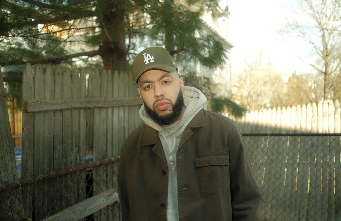 A man with a beard wearing a dark jacket, grey hoodie, and a green Los Angeles Dodgers cap stands outdoors next to a wooden fence and chain-link fence with trees in the background.