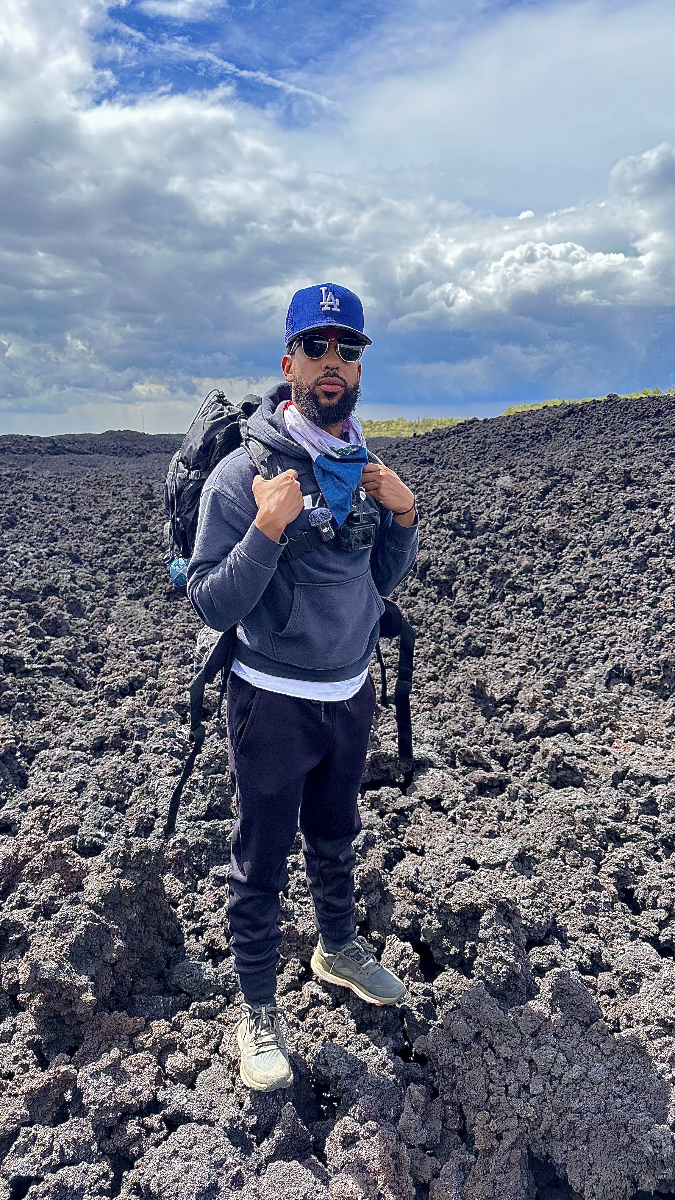 A man wearing a blue Los Angeles Dodgers cap, sunglasses, and hiking gear, standing on rough volcanic terrain with a cloudy sky in the background.