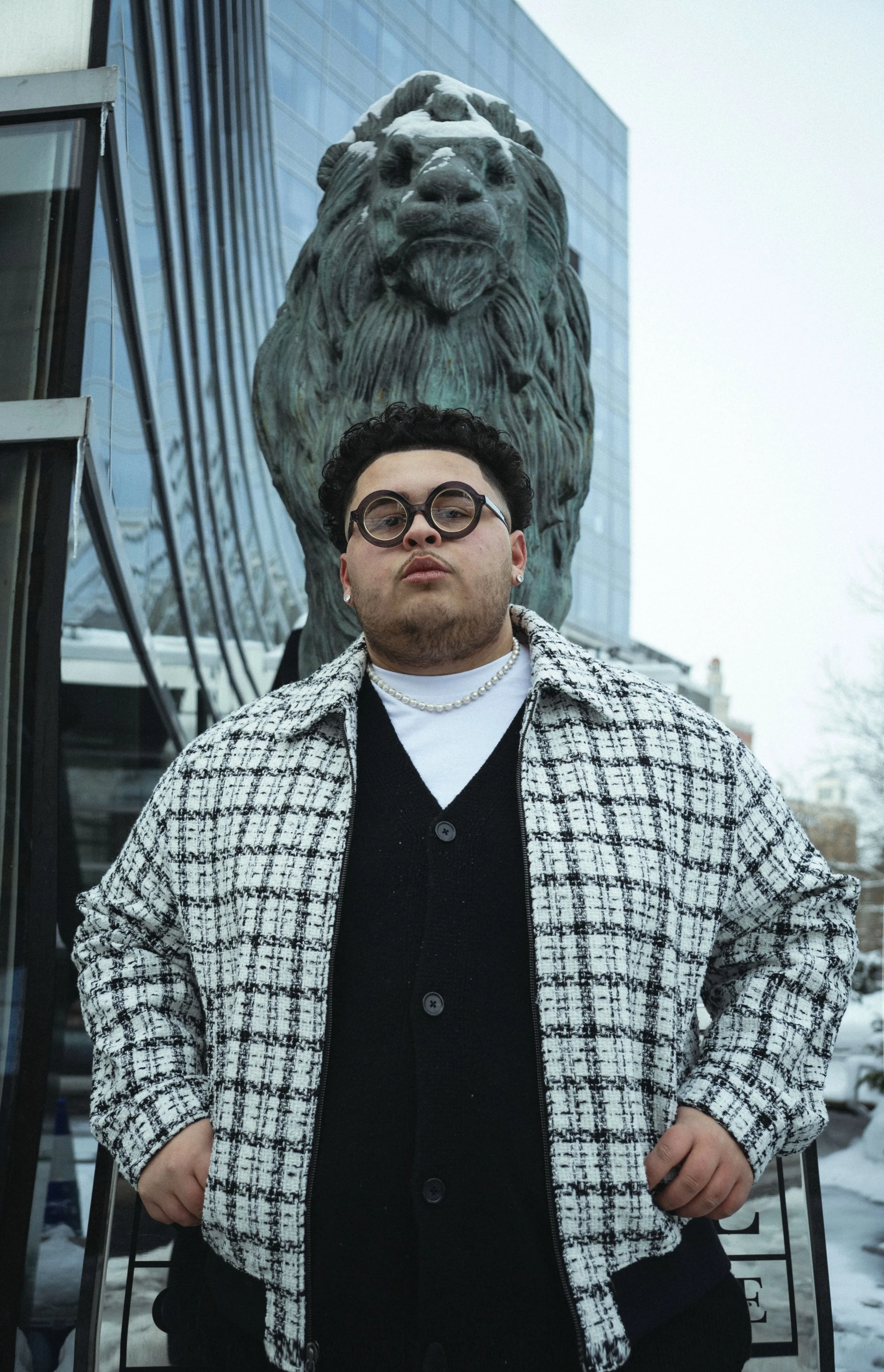 A man wearing round glasses, a pearl necklace, and a checked jacket stands in front of a large lion sculpture on a city street with snow, glass buildings, and trees.