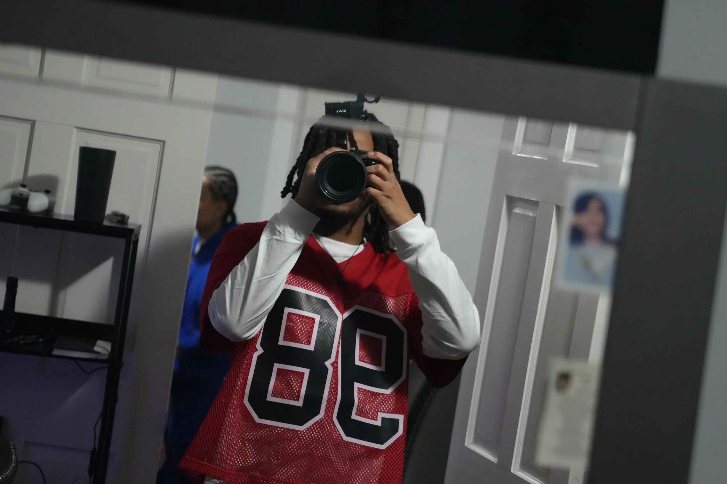 Person taking a photo with a camera in front of a mirror, wearing a red sports jersey with the number 82, in a room with white walls and a door, with a photo of a woman stuck on the mirror.
