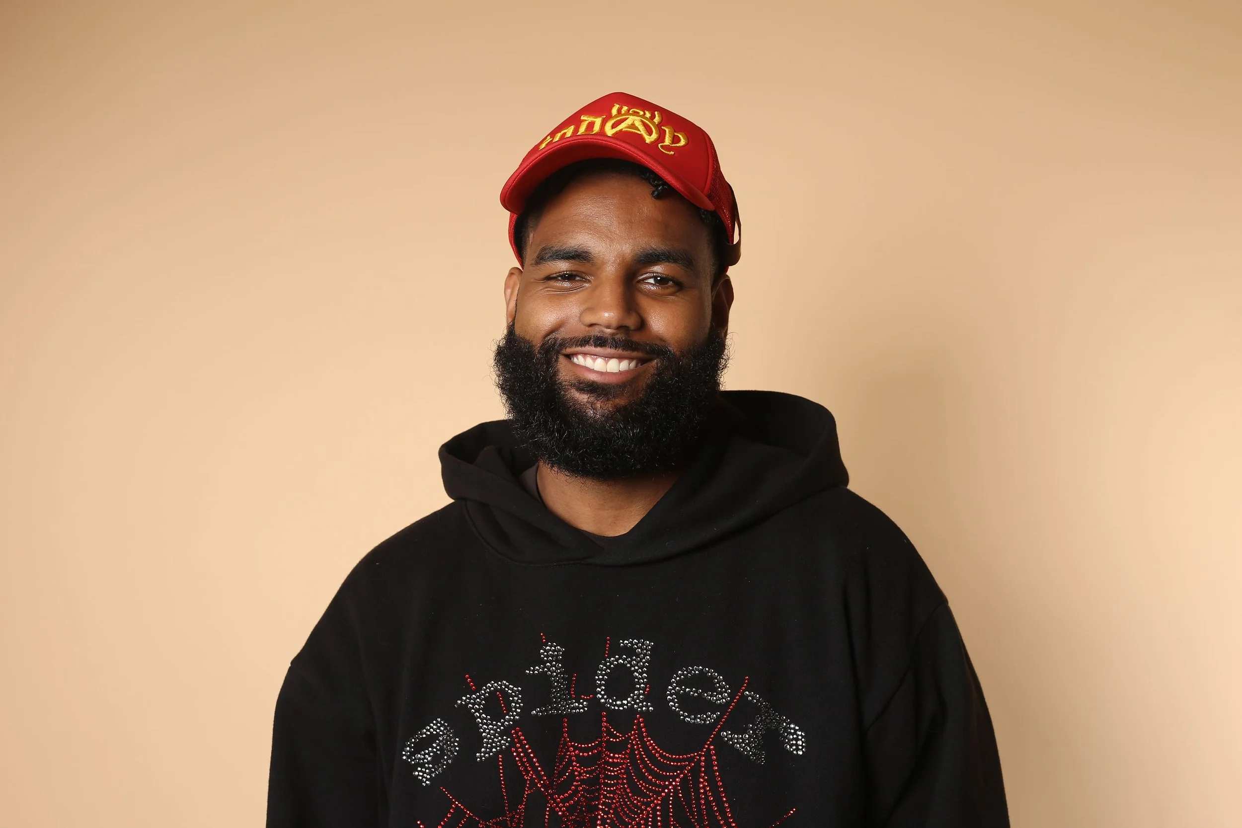 A smiling man with a beard wearing a red cap, black hoodie with a spider web design, standing against a beige background.
