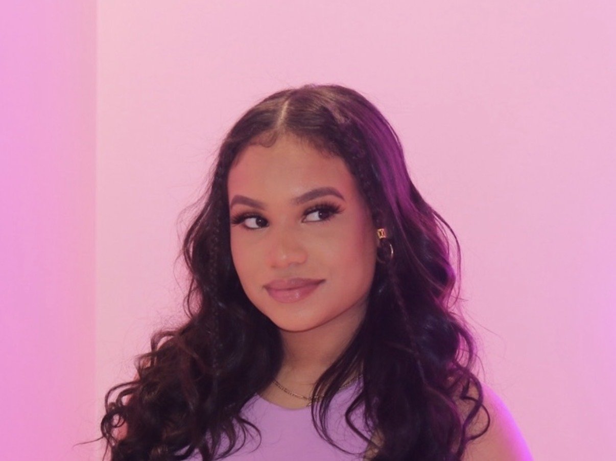 A young woman with long wavy dark hair, wearing a light-colored top and jewelry, smiling softly against a pink background.