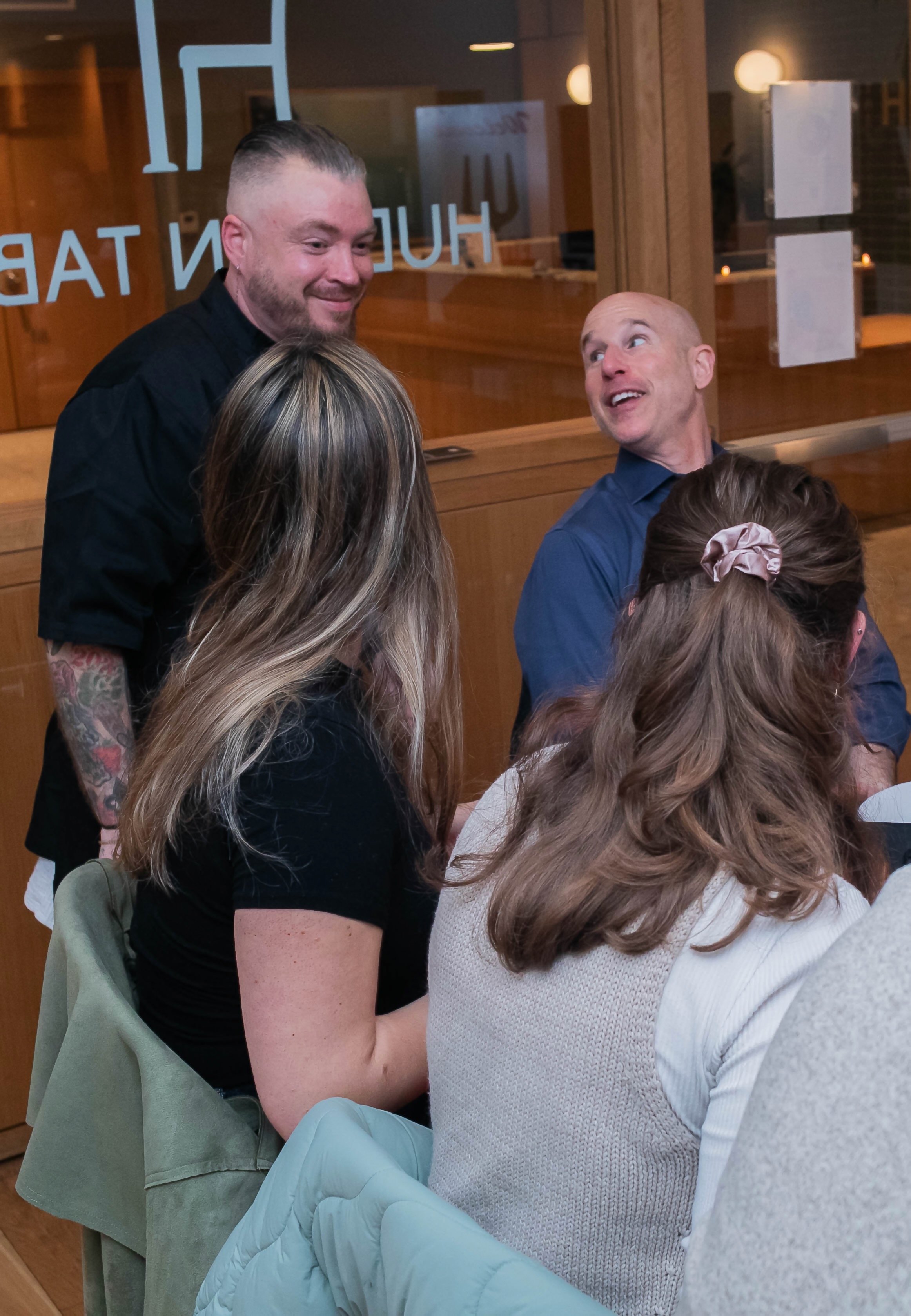 A group of people having a conversation indoors, with one male standing and smiling, while others are seated and looking at him. They appear to be in a modern restaurant or cafe.