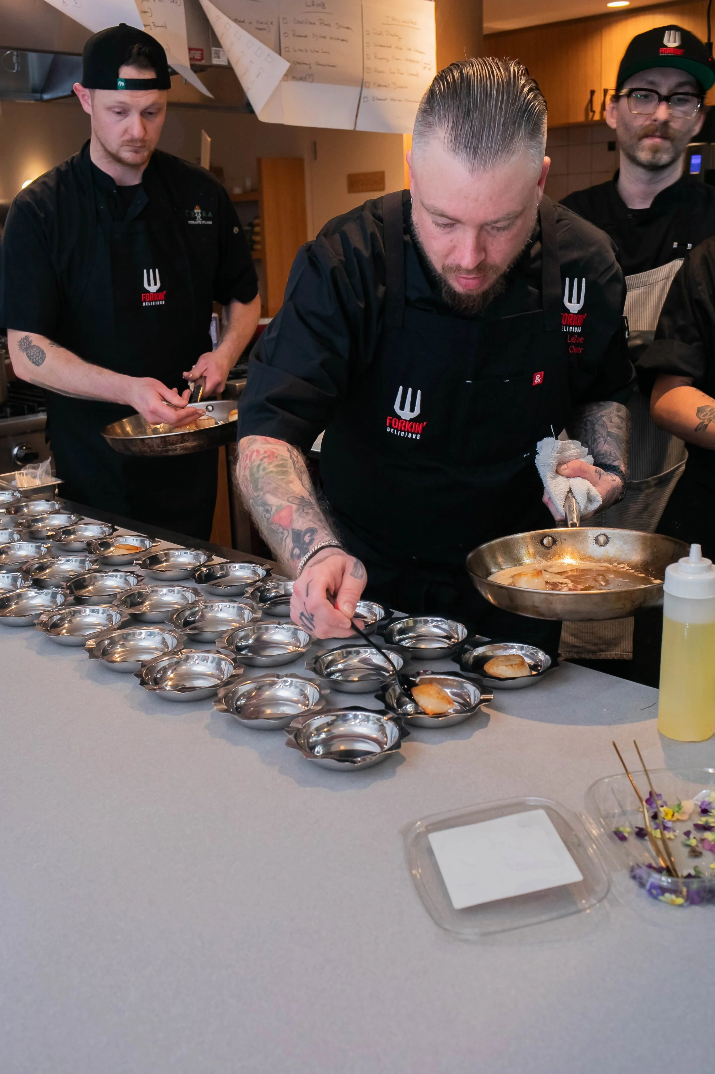 Chefs preparing food in a kitchen, plating dishes in a professional setting, wearing black aprons from Forkin' Delicious.