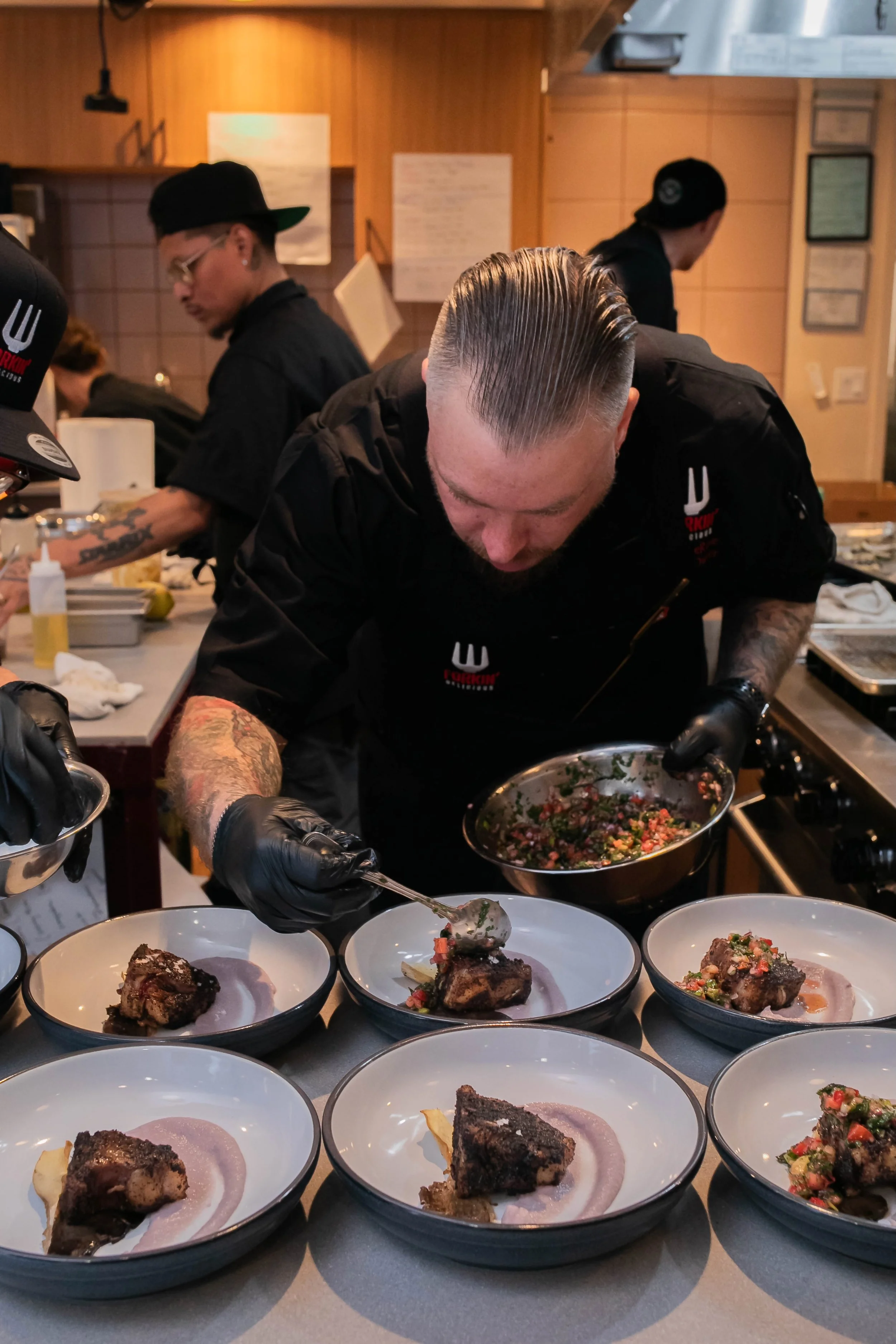 Group of chefs plating dishes with meat and salad garnish in a professional kitchen.