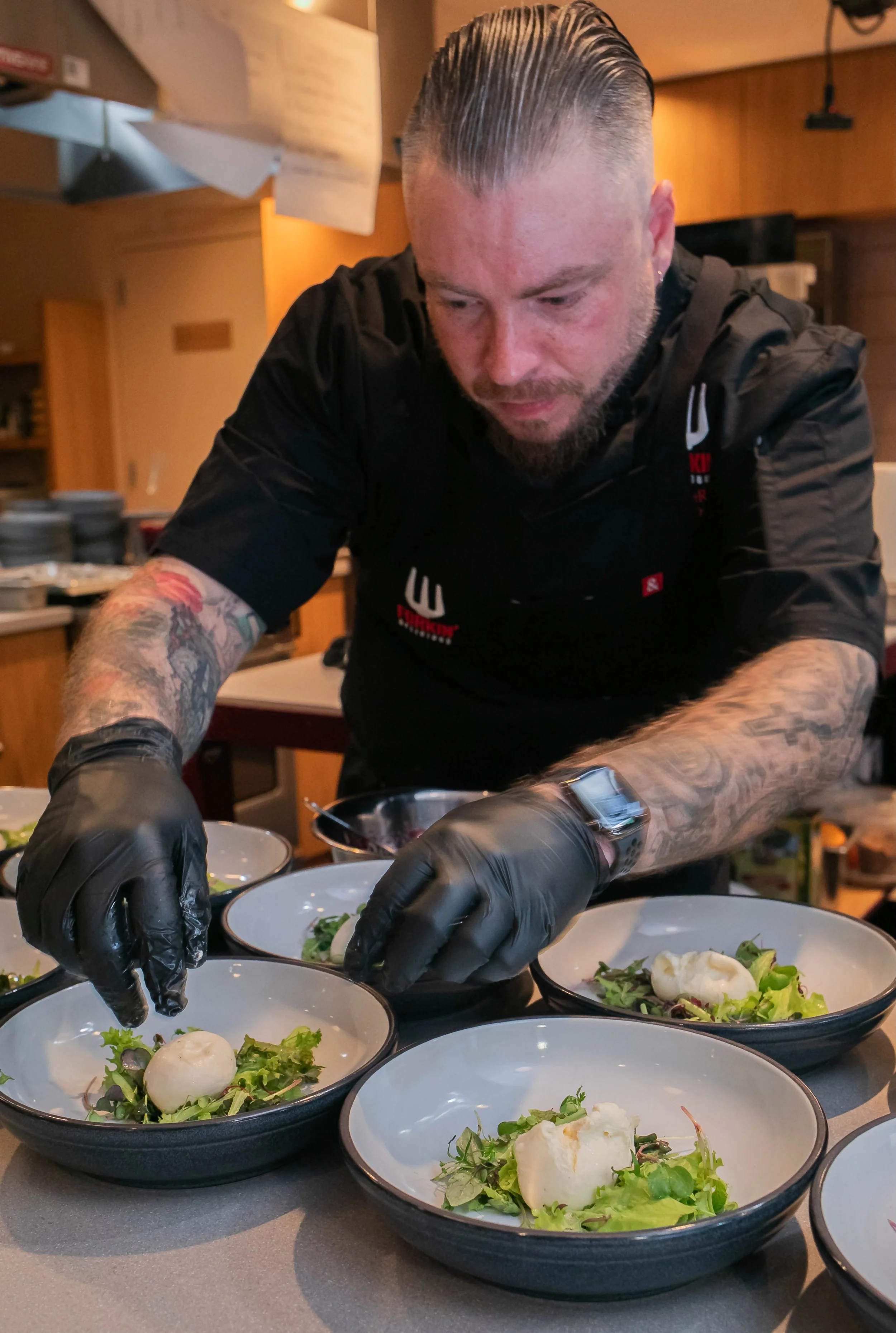 Chef plating dishes in a kitchen, wearing black gloves, with tattoos visible on his arms. Bowls contain mixed greens and a dollop of white mousse.