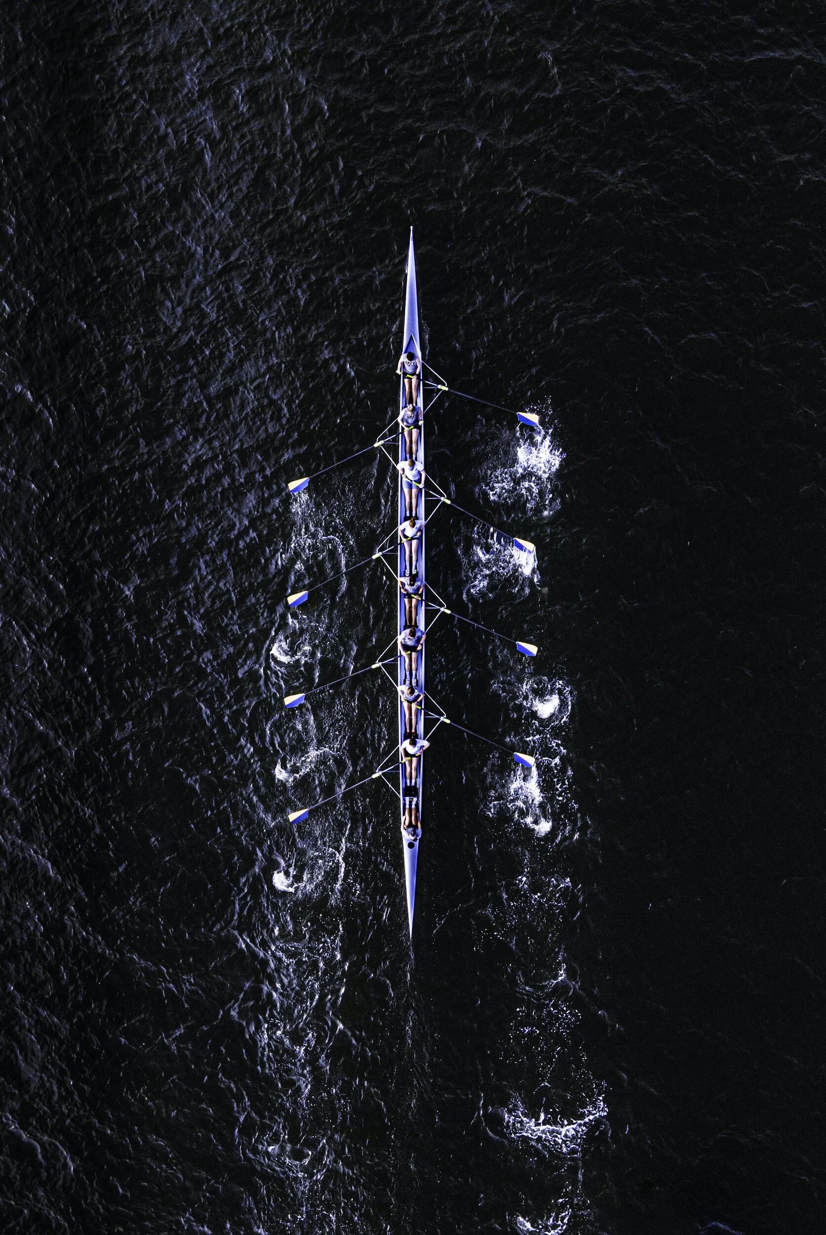 An aerial view of a rowing team racing on a dark body of water, with multiple rowers in a long boat.