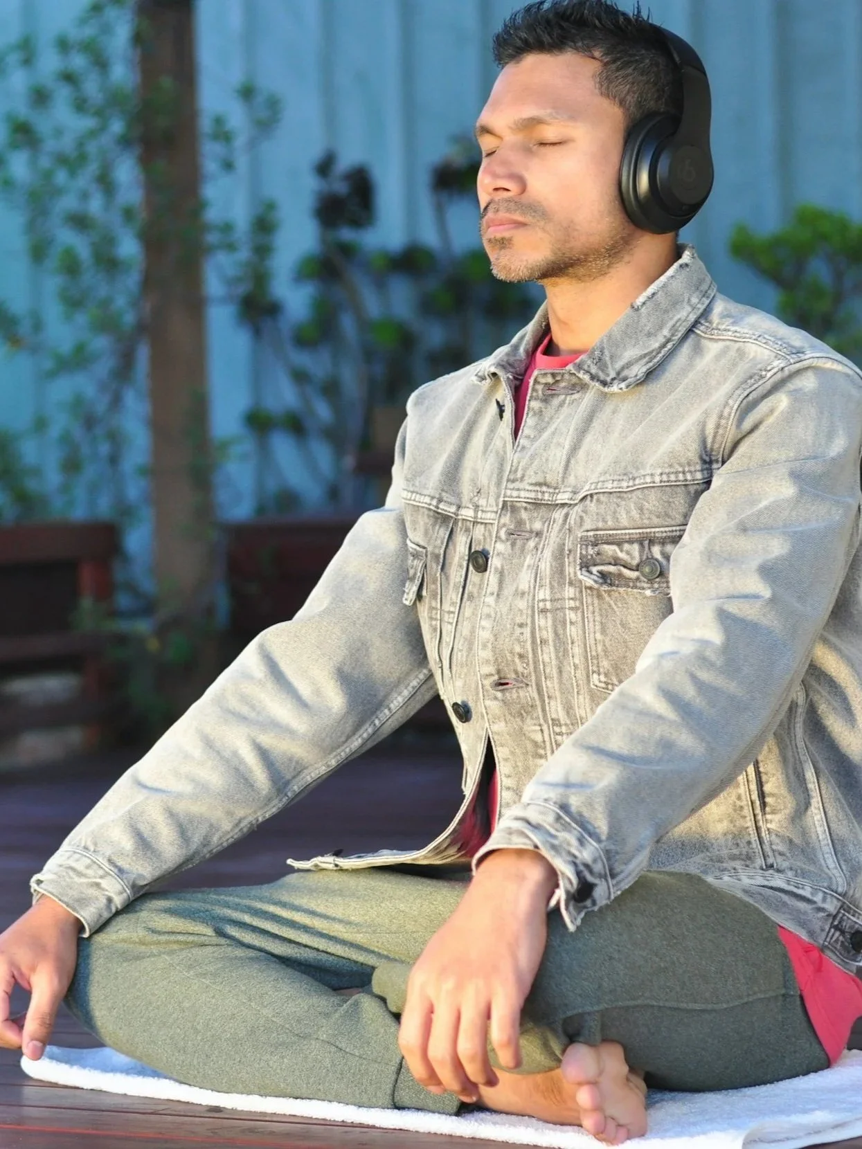 A man meditating outdoors with closed eyes, wearing headphones, a light denim jacket, and green pants, sitting cross-legged on a white mat in a garden setting.