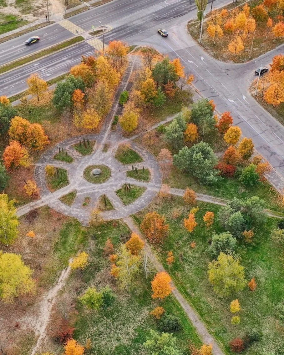 Aerial view of a park with a circular pathway and colorful autumn trees, surrounded by streets with moving cars.
