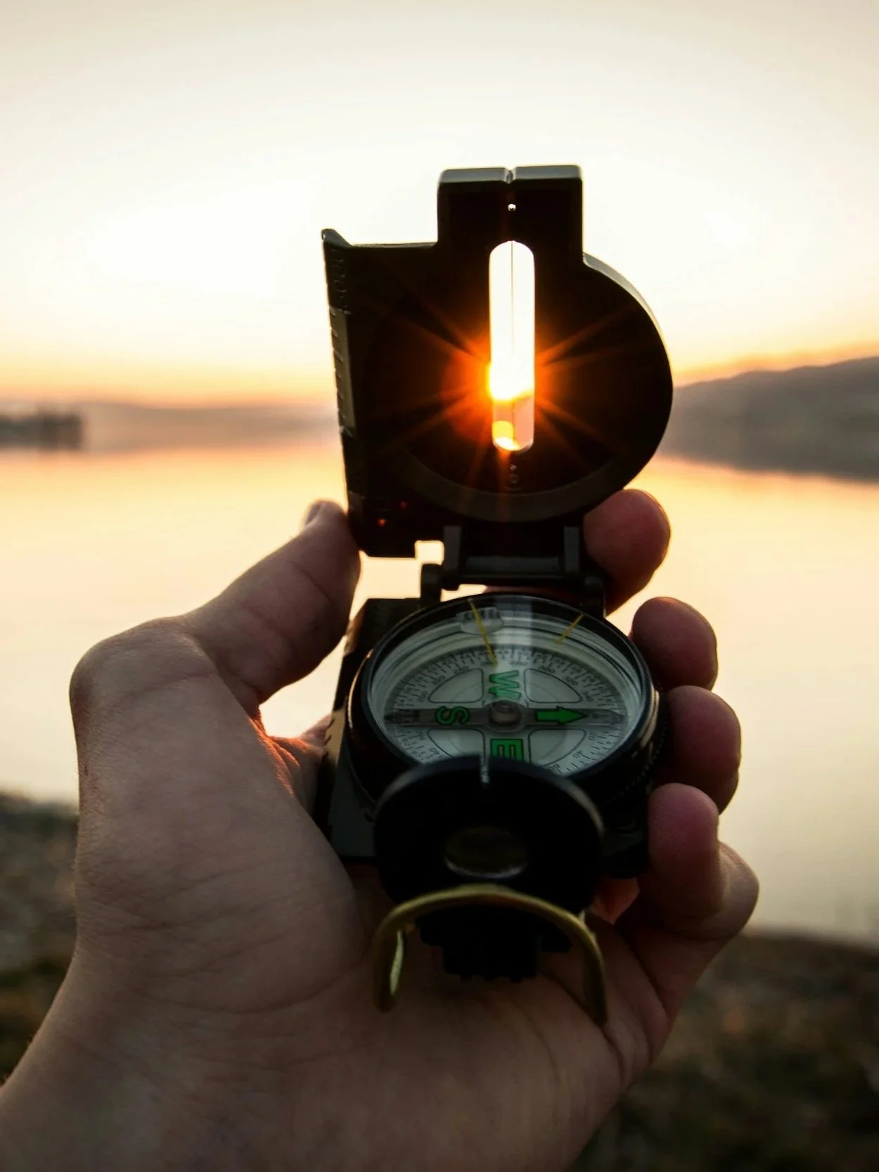 A person holds a compass with a sun setting behind it, creating a bright glow through a lens at the top of the compass, with a lake and a distant shoreline in the background.