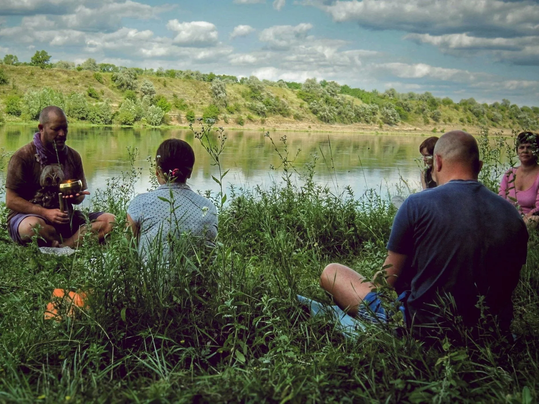 People sitting by a river, engaging in a meditation activity, surrounded by greenery and a cloudy sky.