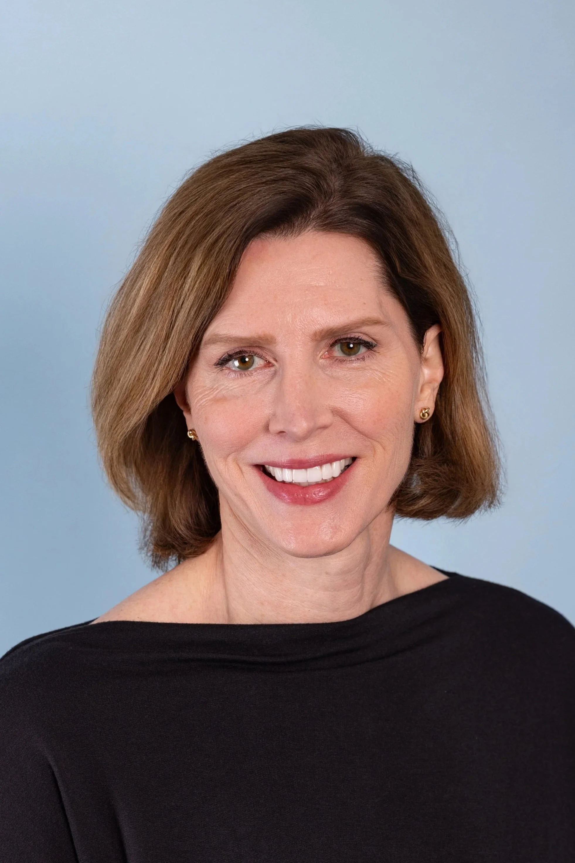 Close-up of a smiling woman with shoulder-length brown hair wearing a black top against a light blue background.
