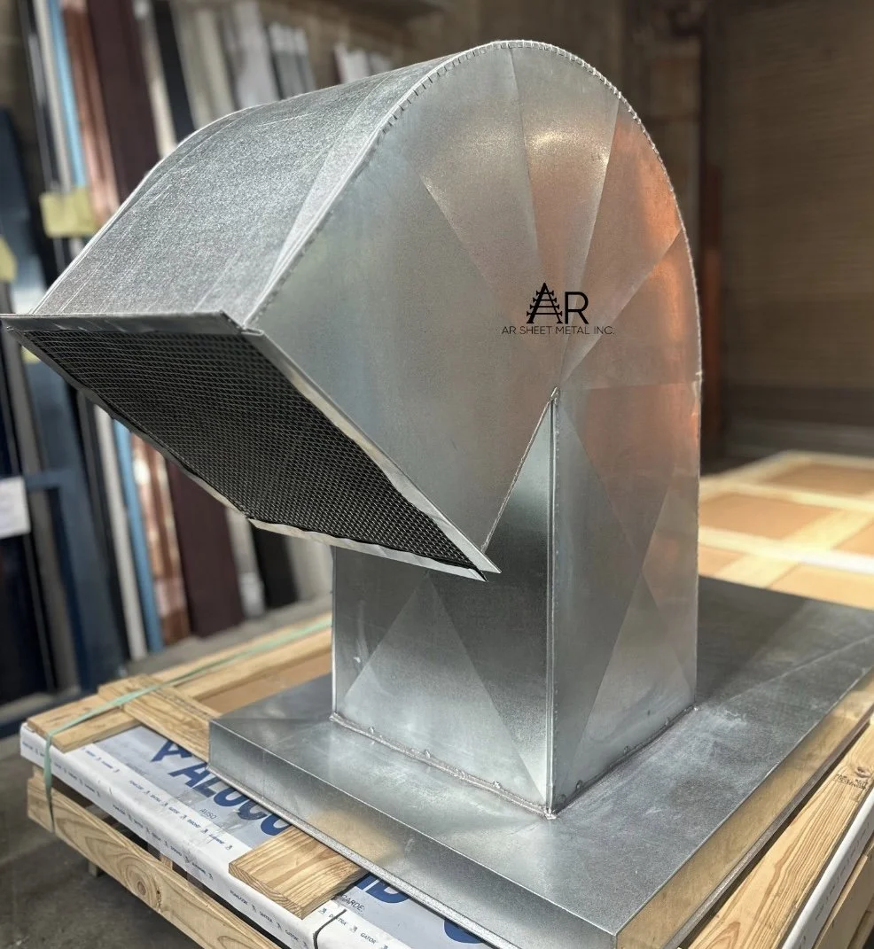 A close-up view of a metallic HVAC duct component with an angled vent, mounted on a wooden platform with building materials in the background.