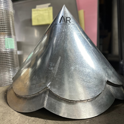 Metal baking mold in the shape of a heart with a pointed top, placed on a table with some stacked metallic circular objects and a pink background in the background.