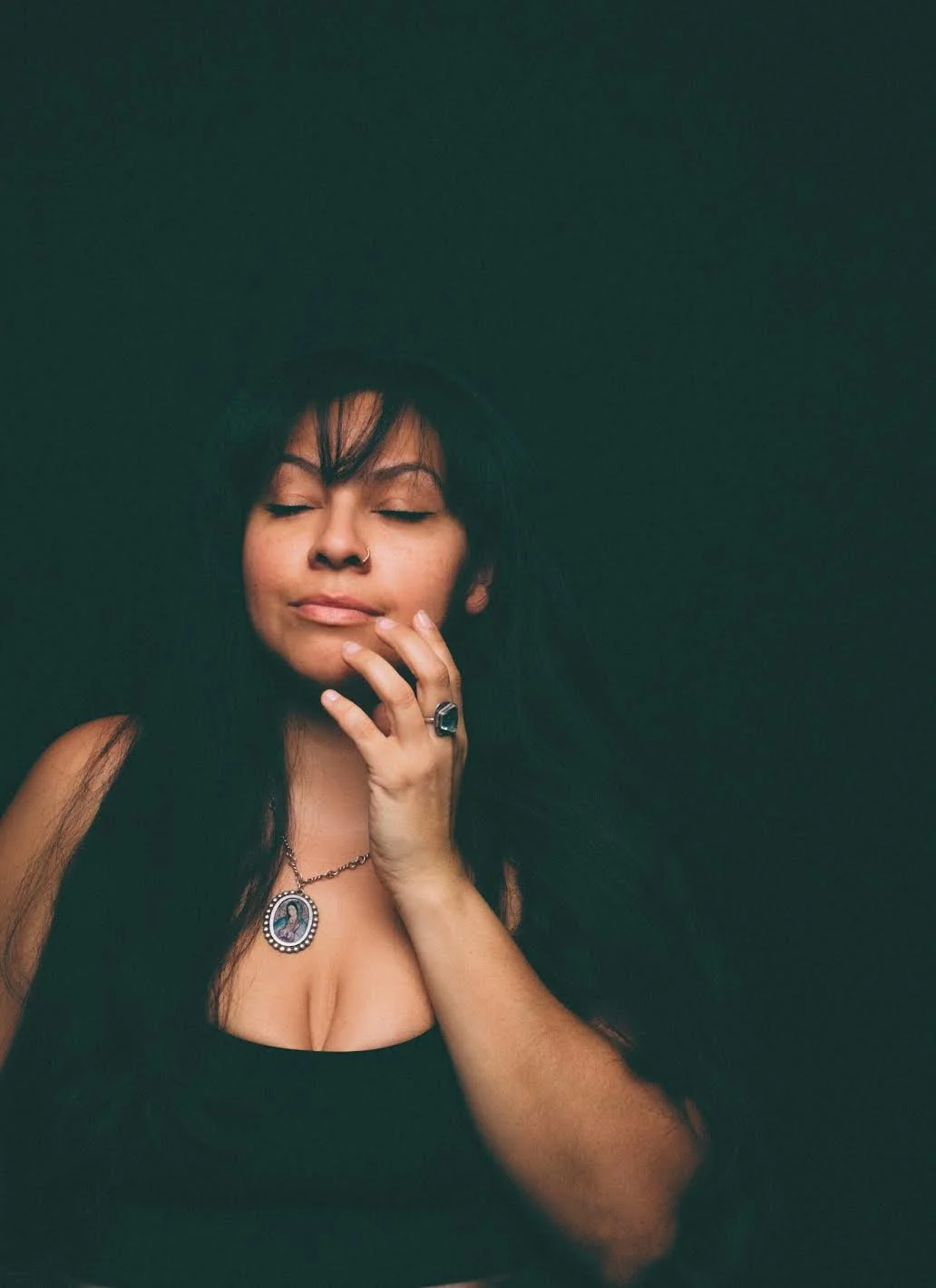 A woman with dark hair, closed eyes, wearing a black tank top, a hat, a ring, a necklace with a religious pendant, and a nose ring, touching her face with one hand against a plain dark background.