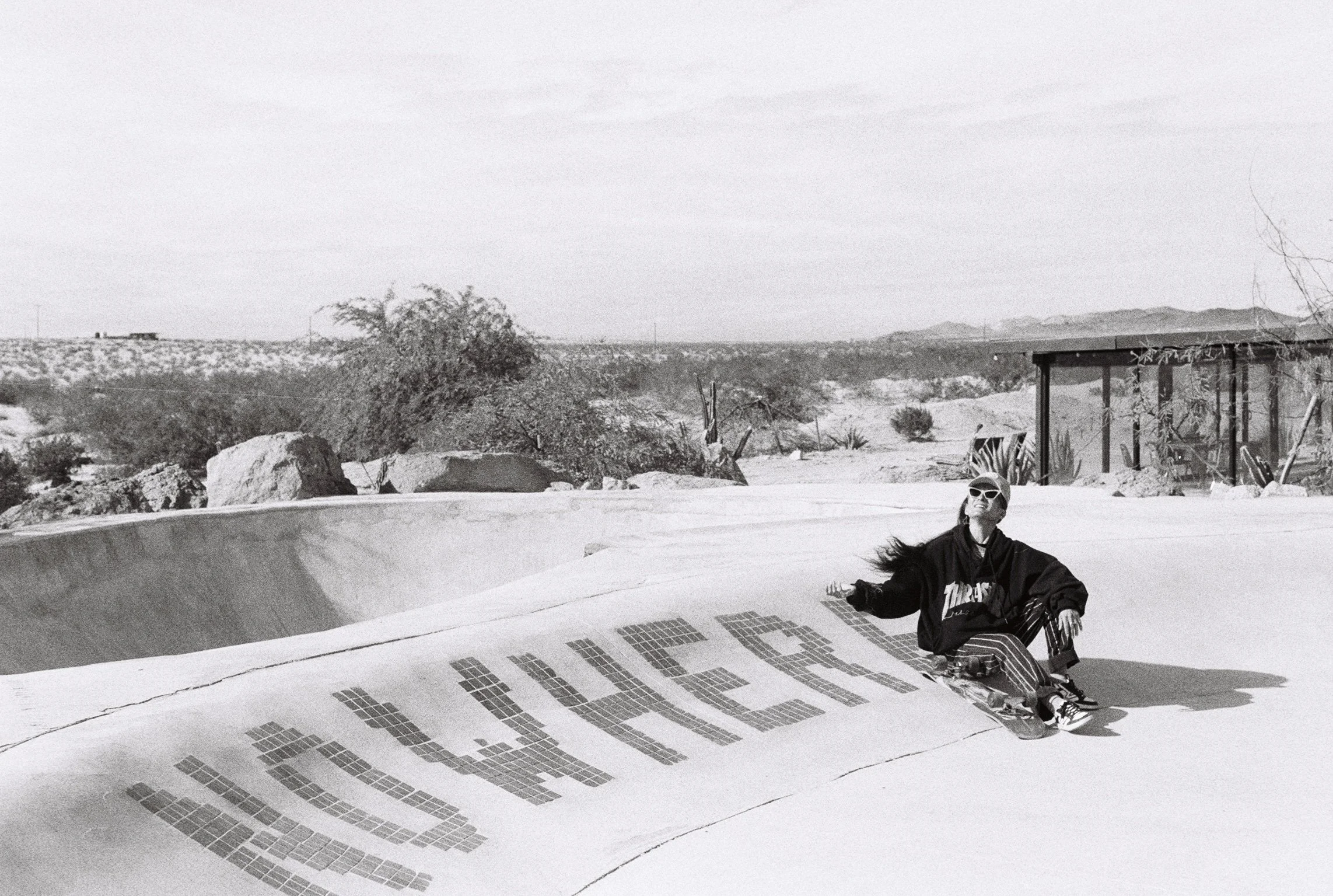 Person sitting on a skateboard in a desert landscape wearing sunglasses and a dark hoodie, in black and white.