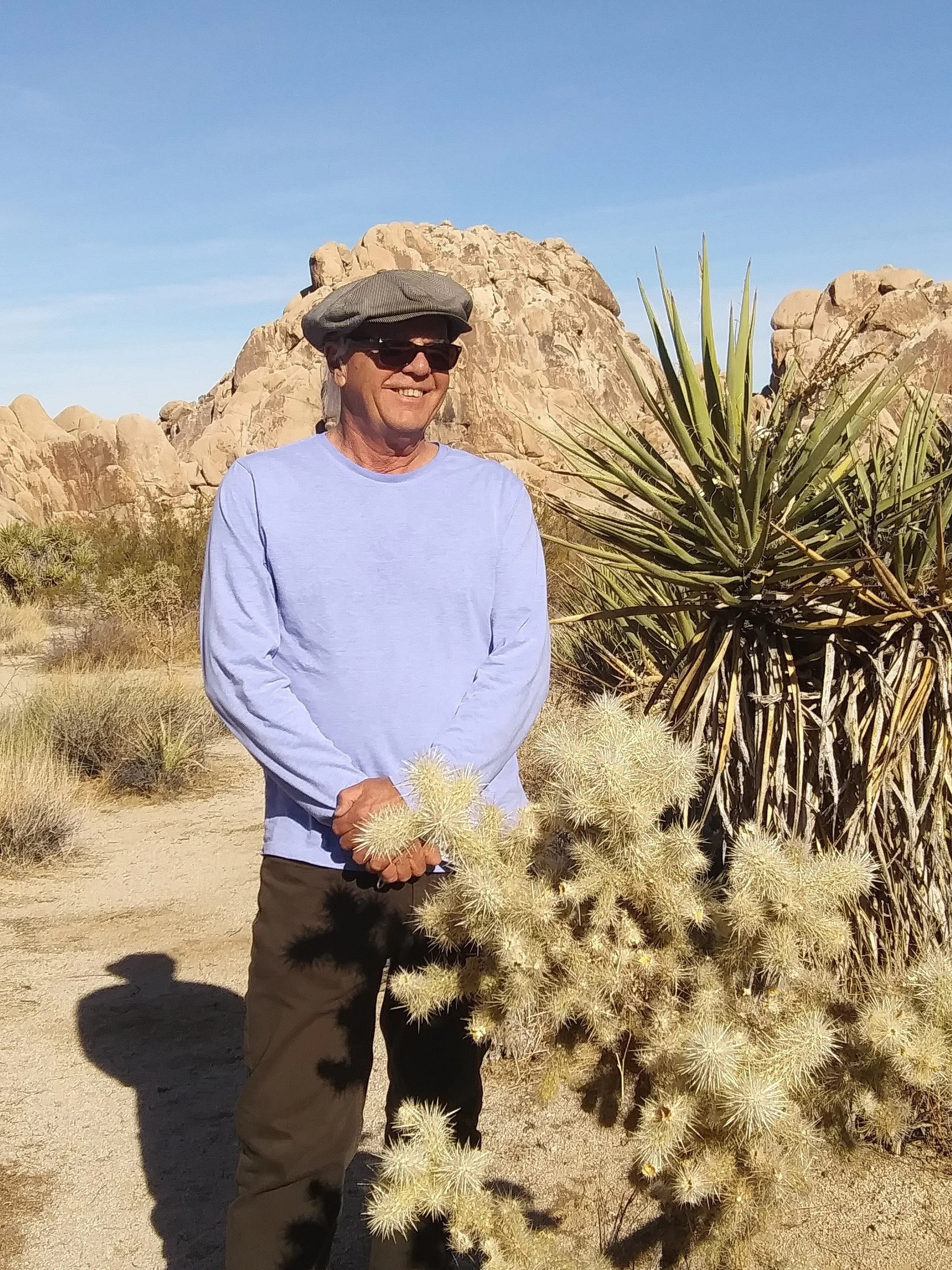 A smiling man wearing sunglasses, a gray hat, and a light blue shirt standing in a desert landscape with cacti and rocky formations.