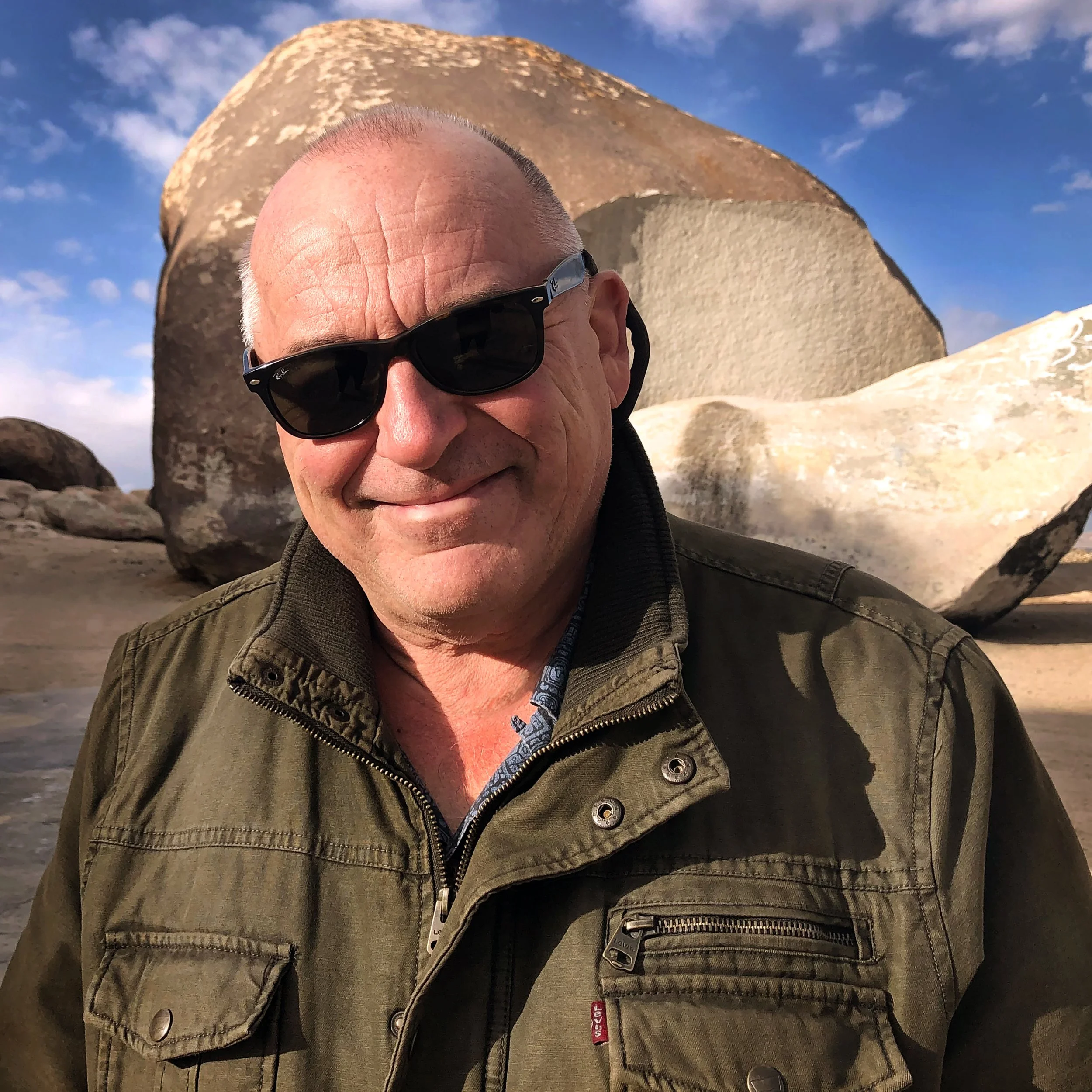 A smiling man with sunglasses and a green jacket standing on a beach with large boulders in the background and a partly cloudy sky.