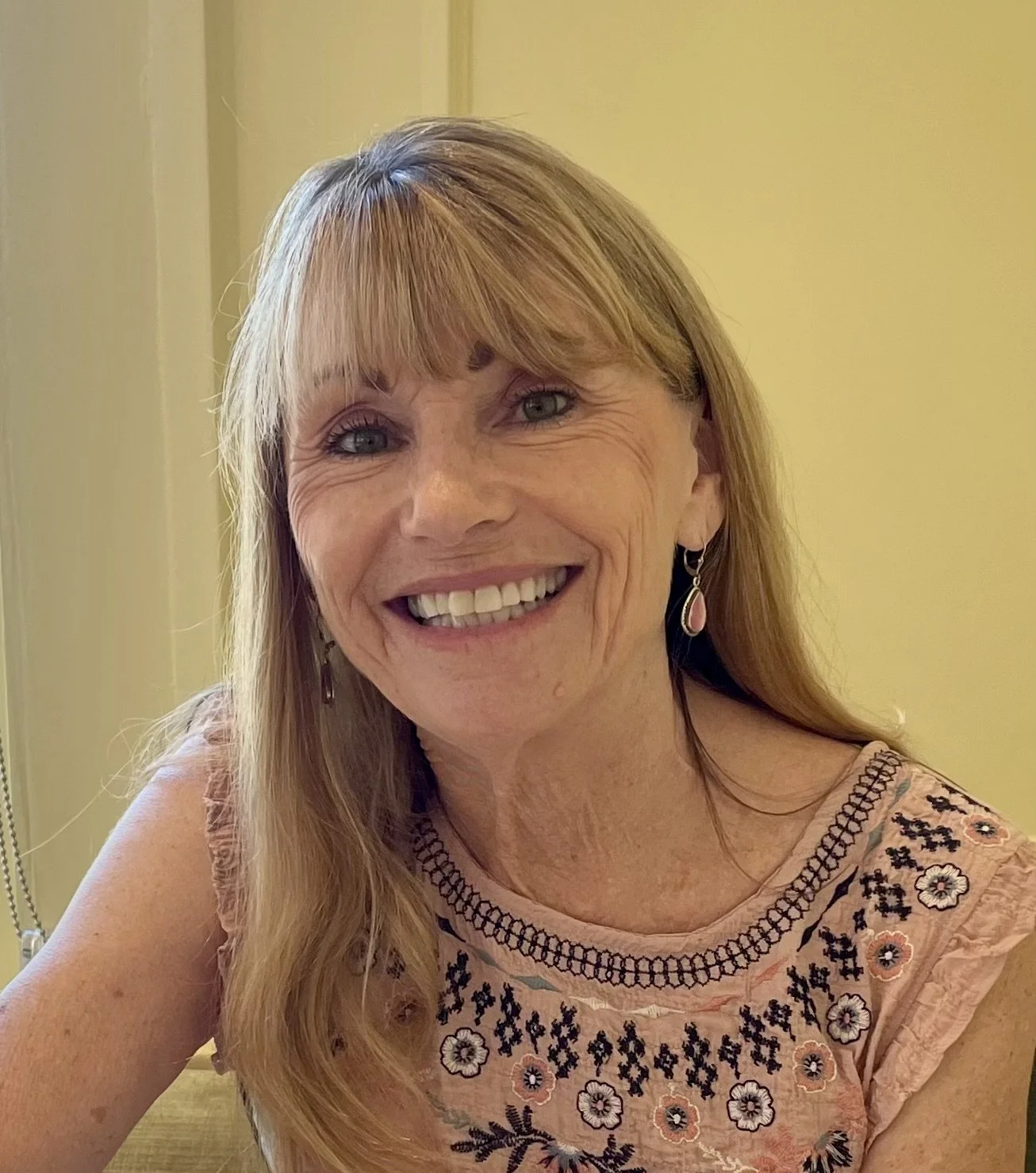 A smiling woman with long strawberry blonde hair, wearing a pink embroidered top and teardrop earrings, sitting indoors against a yellow wall.