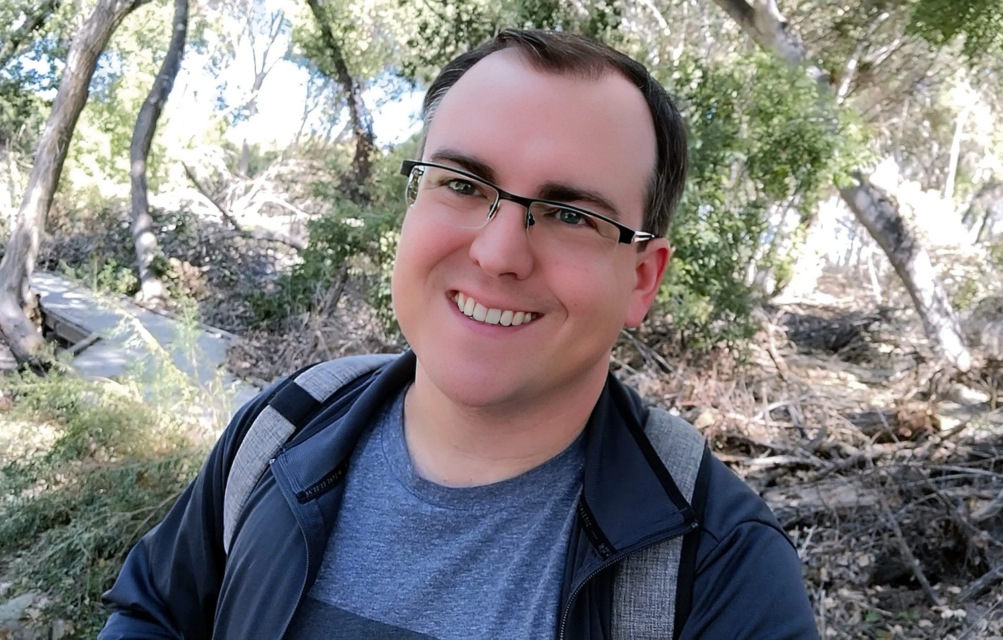 A smiling man with glasses taking a selfie outdoors in a forested area.