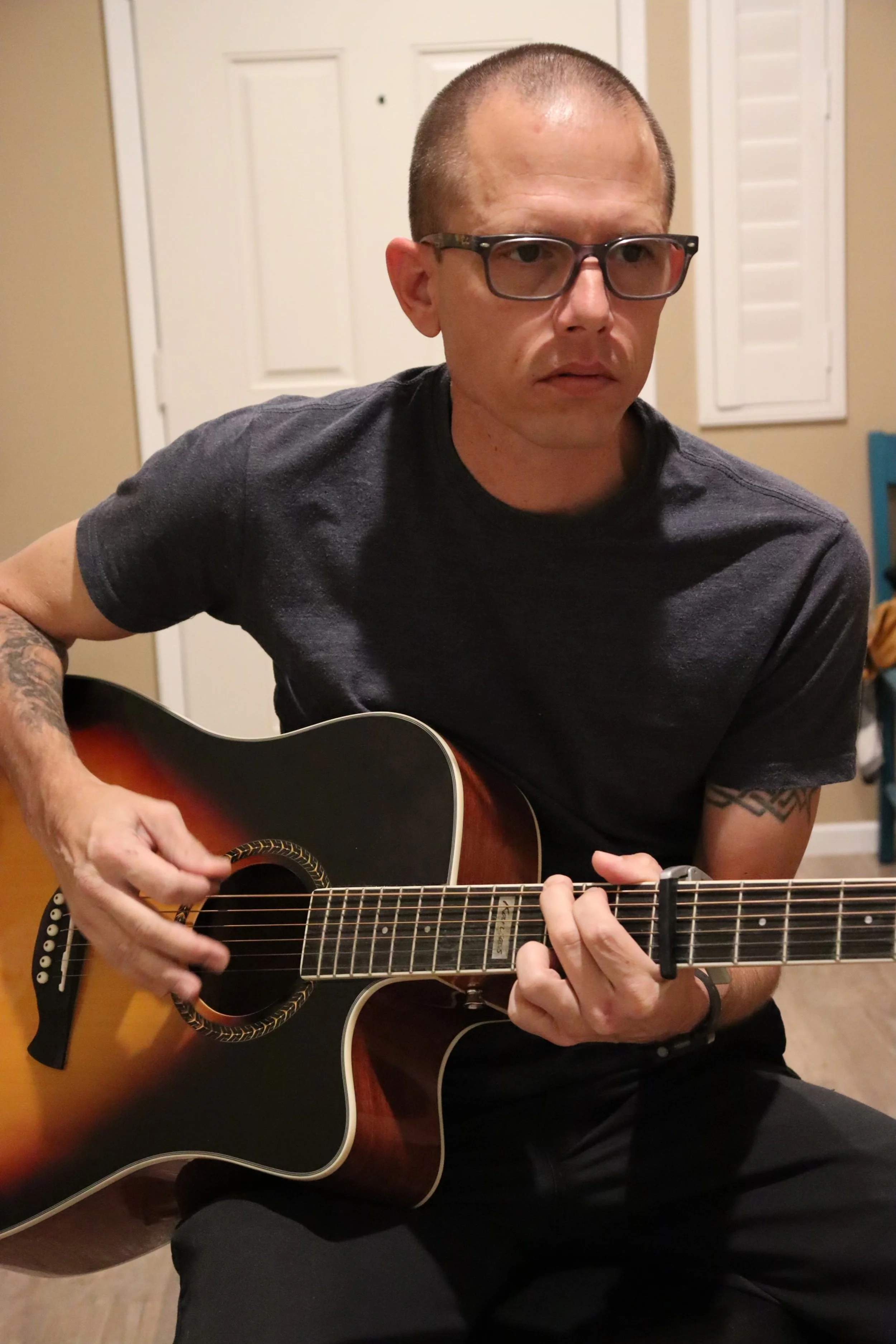 A man with short hair, glasses, and tattoos playing an acoustic guitar indoors.