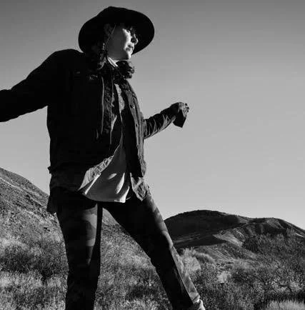 Person wearing a hat and jacket standing outdoors in a hilly landscape.