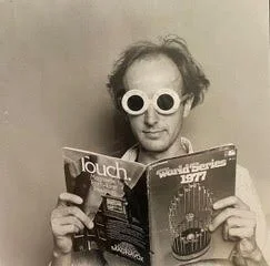 A man wearing round glasses reading a magazine called 'Touch' against a plain background.