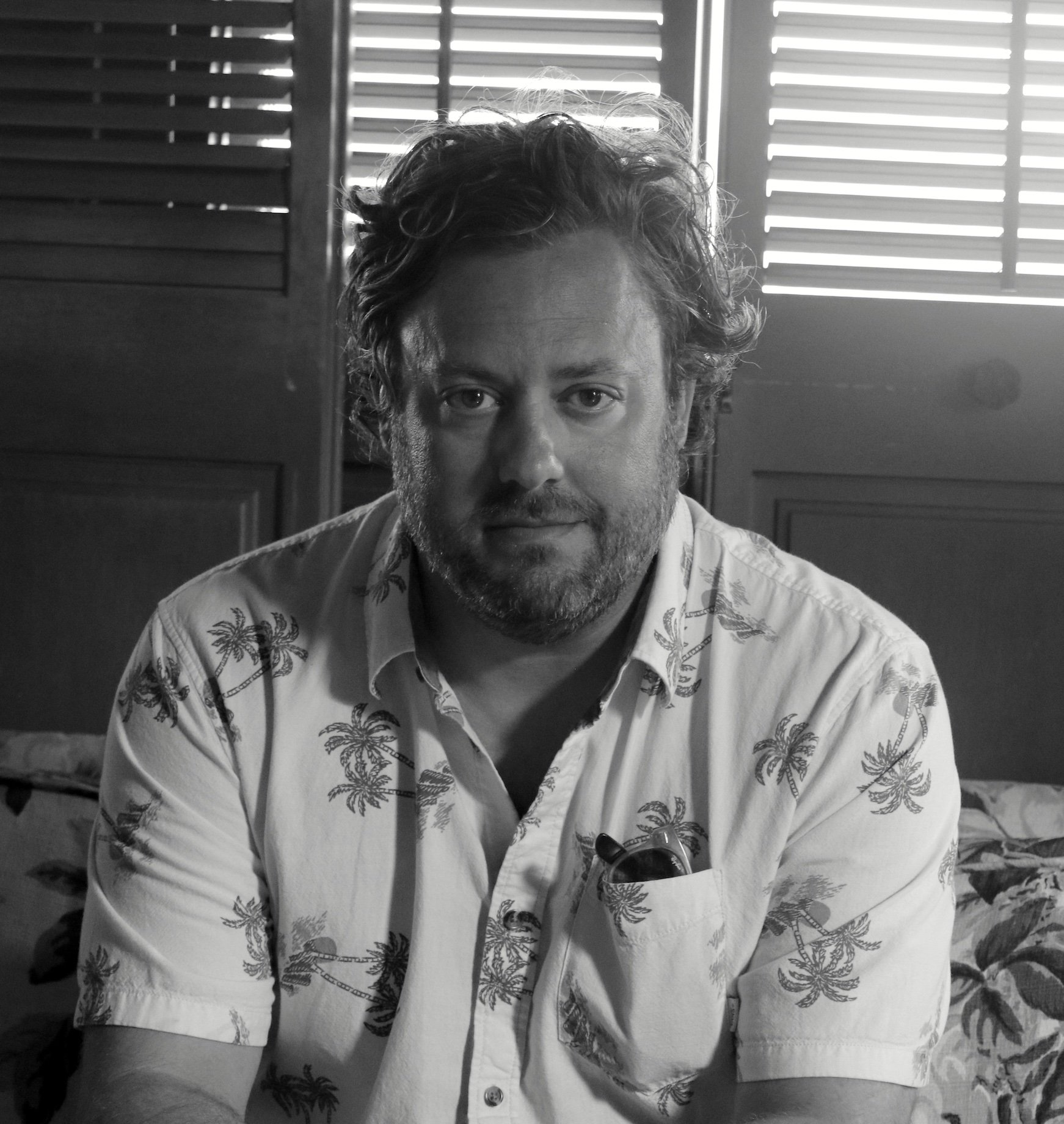 A man with curly hair and a beard sitting indoors, wearing a Hawaiian shirt with palm trees, in black and white photograph.