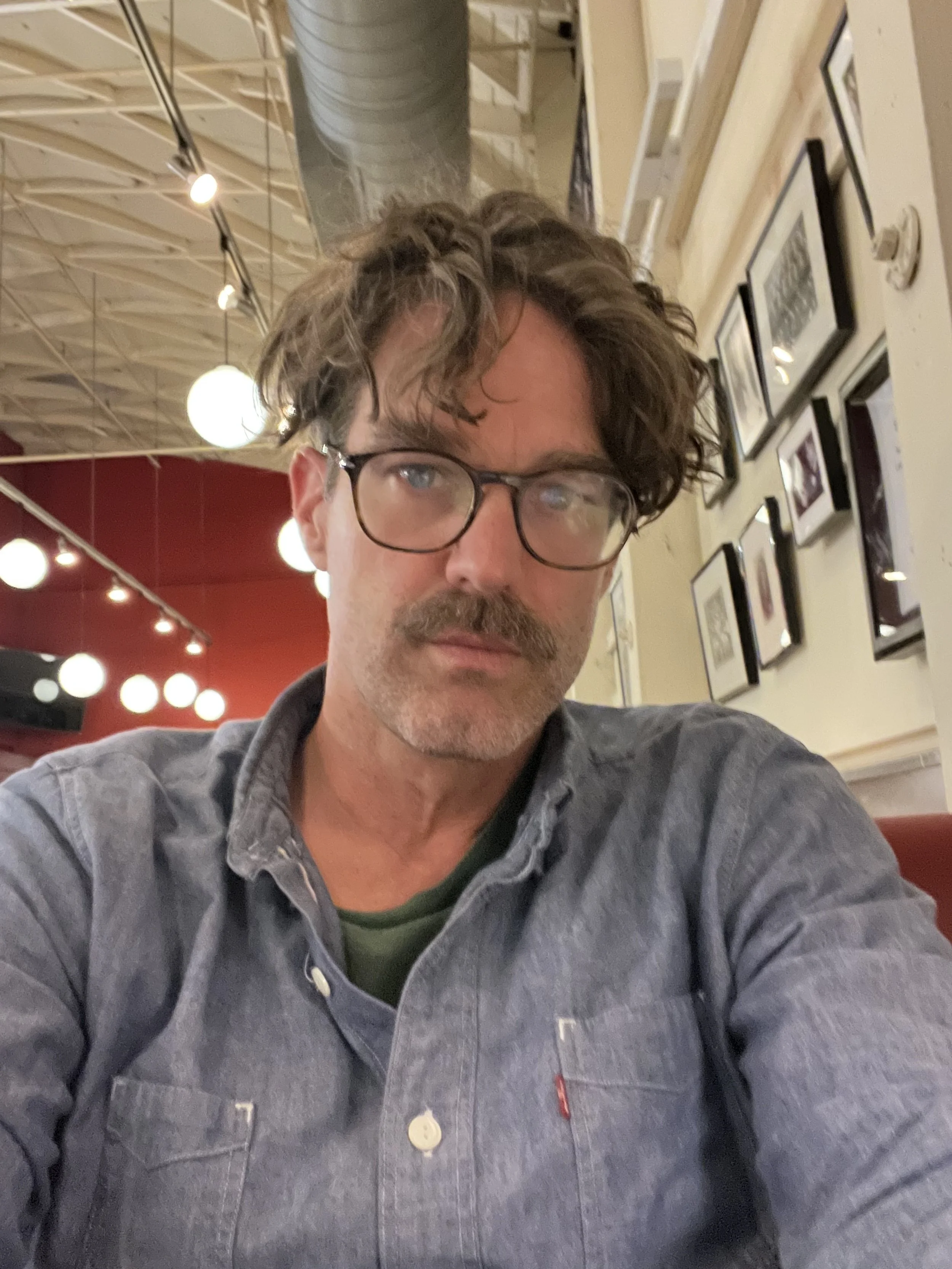 A man with wavy dark hair, glasses, and a mustache taking a selfie in a restaurant or cafe with framed pictures on the wall and warm lighting.