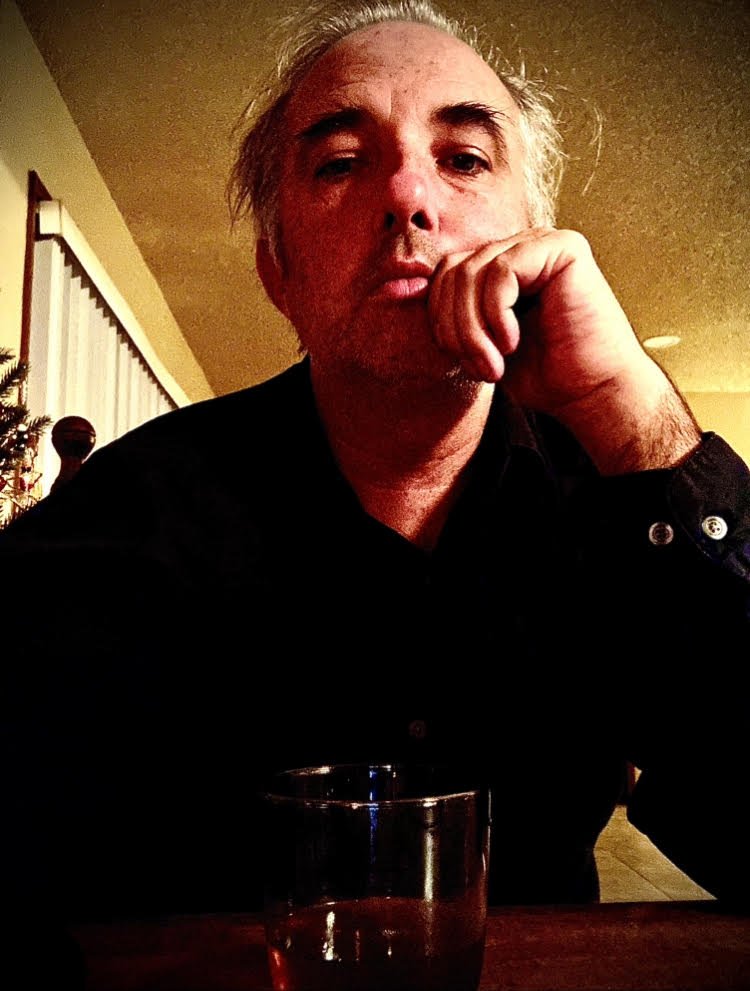 A man with gray hair sitting at a table with a glass of dark-colored beverage. He is resting his chin on his hand and looking at the camera.