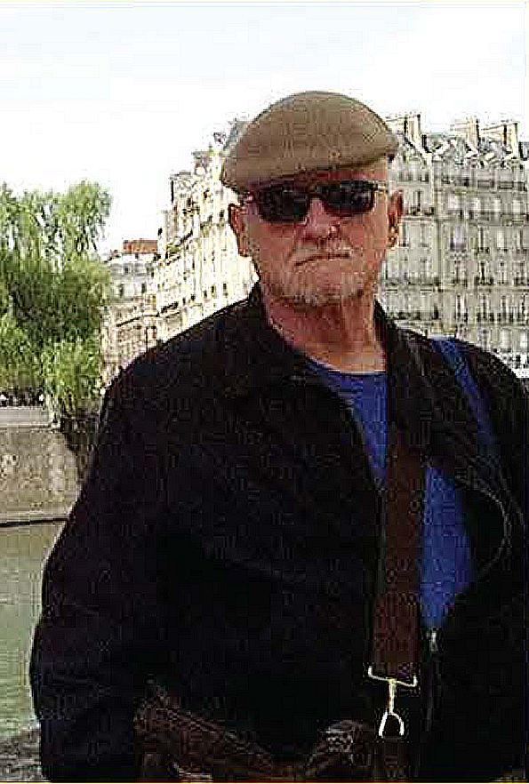 Image of a man wearing sunglasses, a beige flat cap, and a black jacket, standing outdoors in front of classic European-style buildings with a canal on the left side.