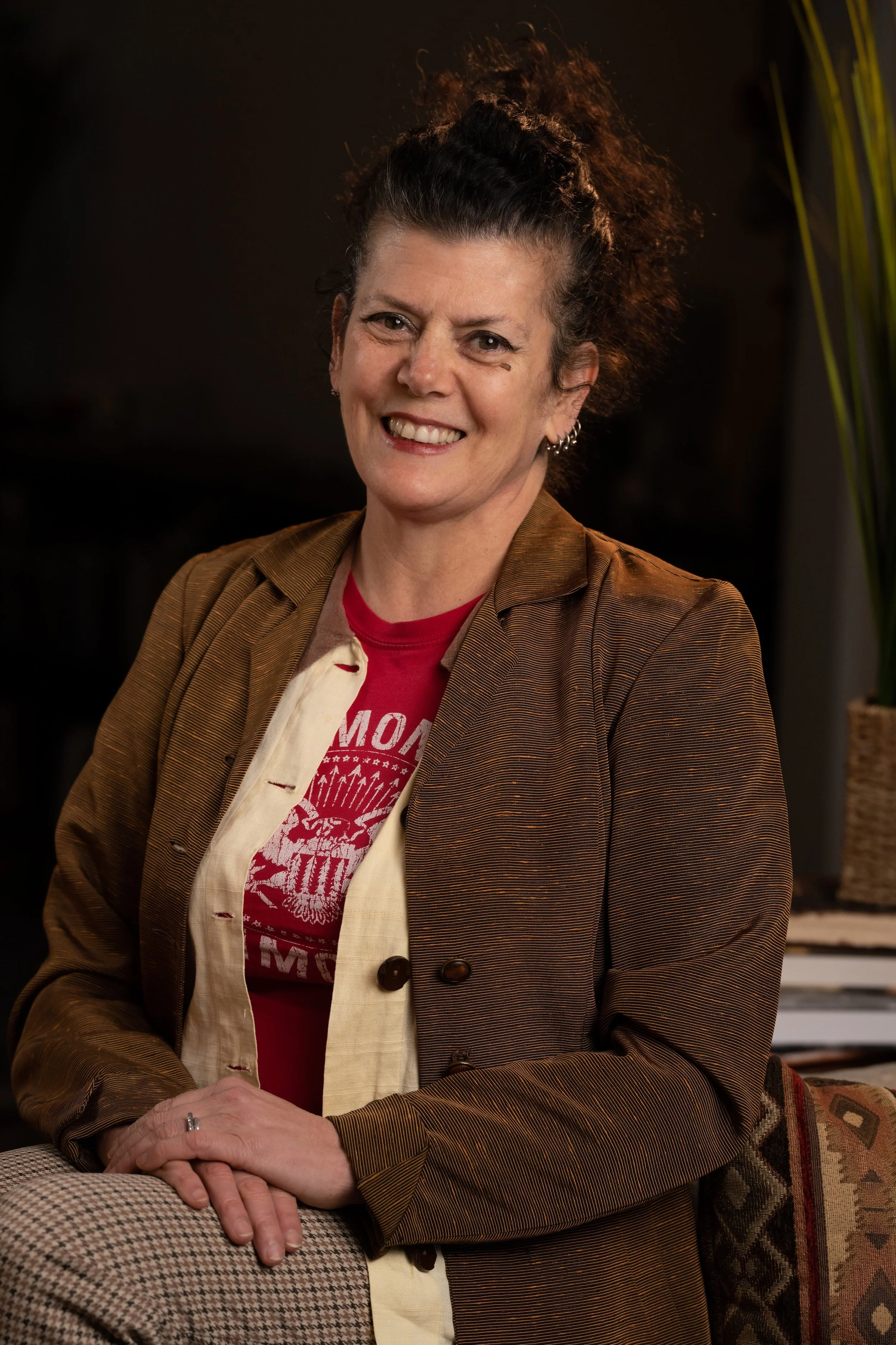 A woman with curly dark hair smiling, wearing a brown jacket and a red graphic T-shirt, sitting with her hands clasped, in an indoor setting.