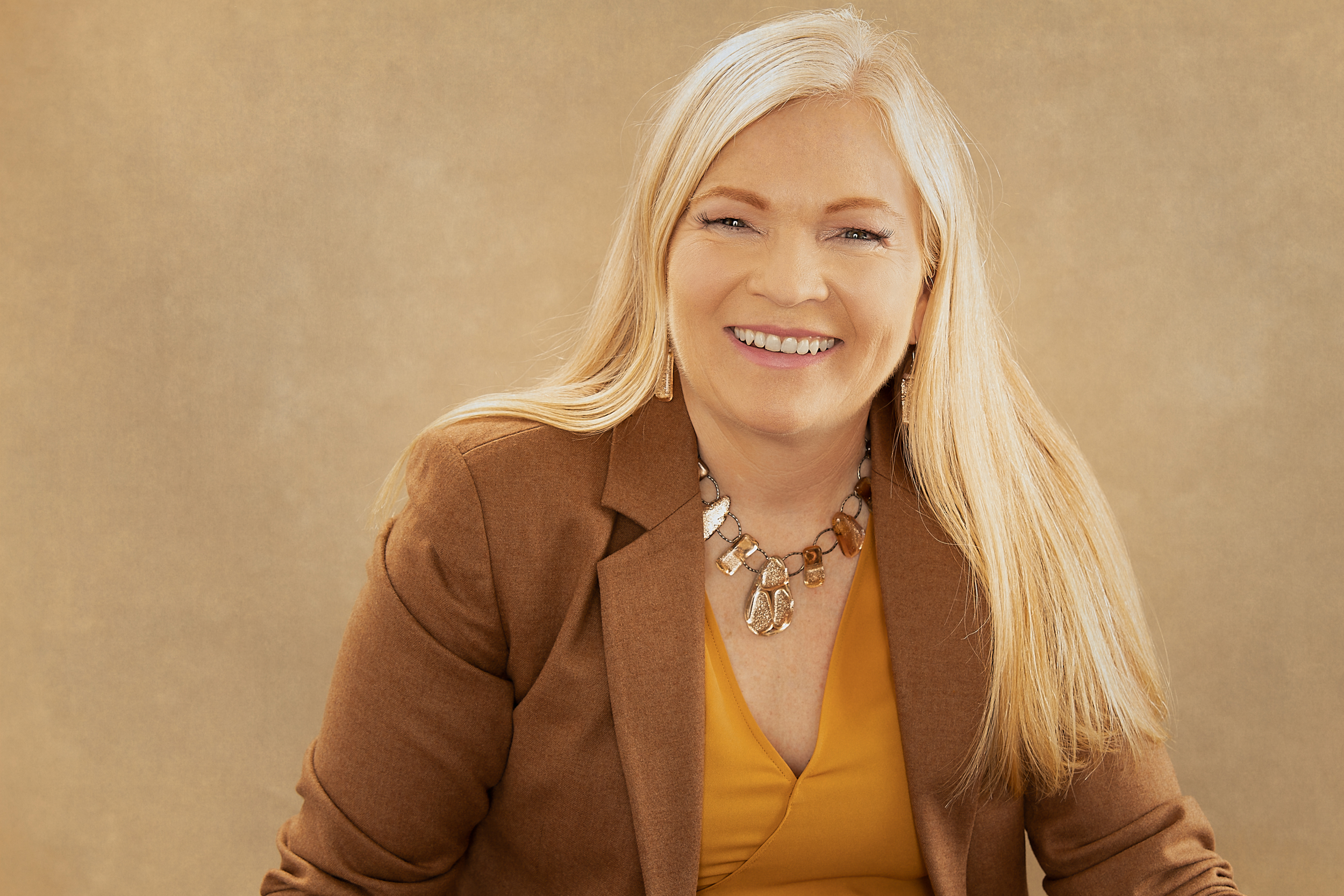 A woman with long blonde hair, smiling, wearing a brown blazer over a yellow top with a statement necklace, against a plain beige background.