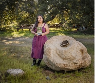 A woman in a purple dress standing outdoors next to a large rock with bowls on it on a grassy area surrounded by trees.