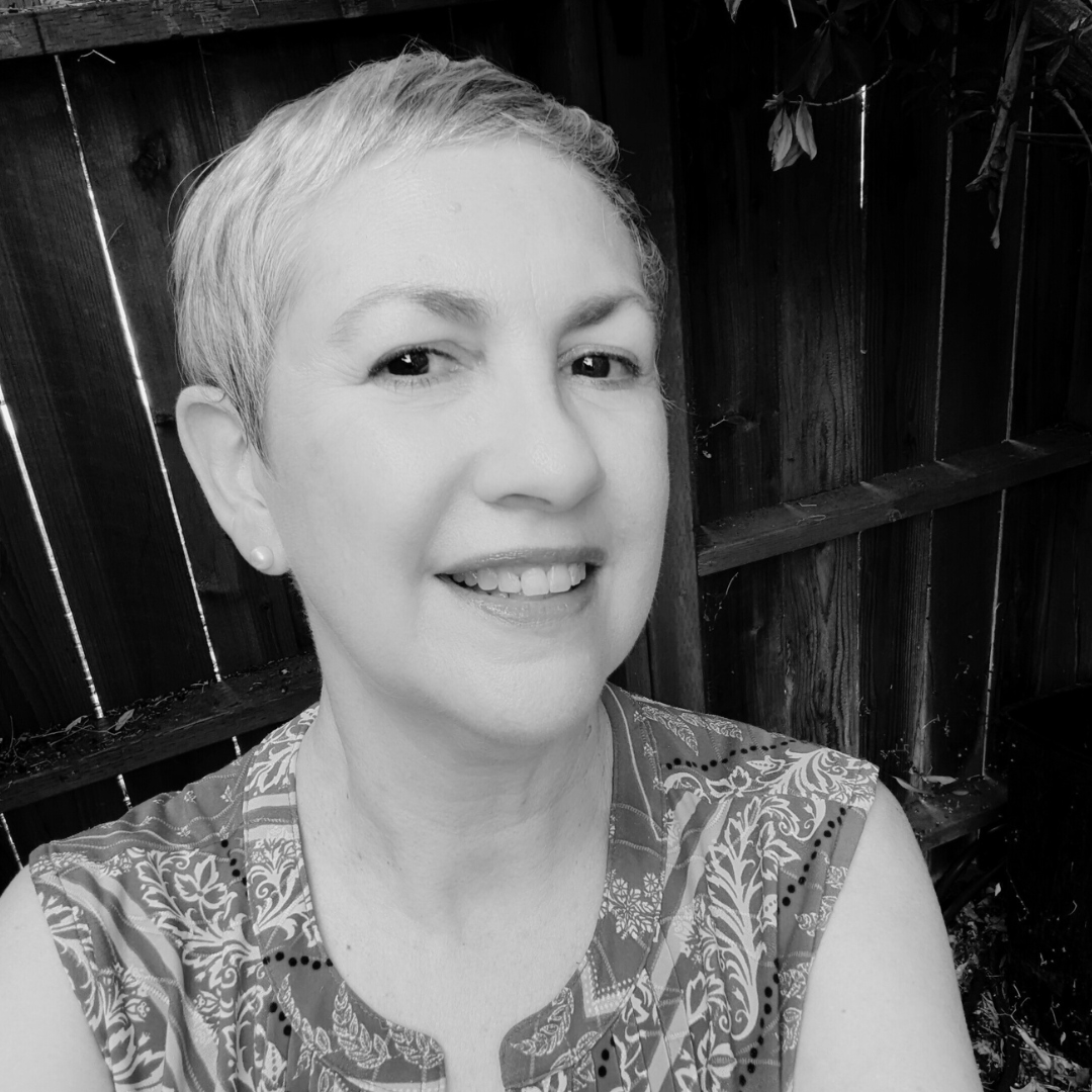 A smiling woman with short hair taking a selfie outdoors against a wooden fence.