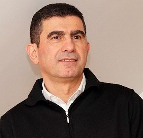 A man with short dark hair wearing a black zip-up jacket over a white collared shirt, looking slightly to the side.